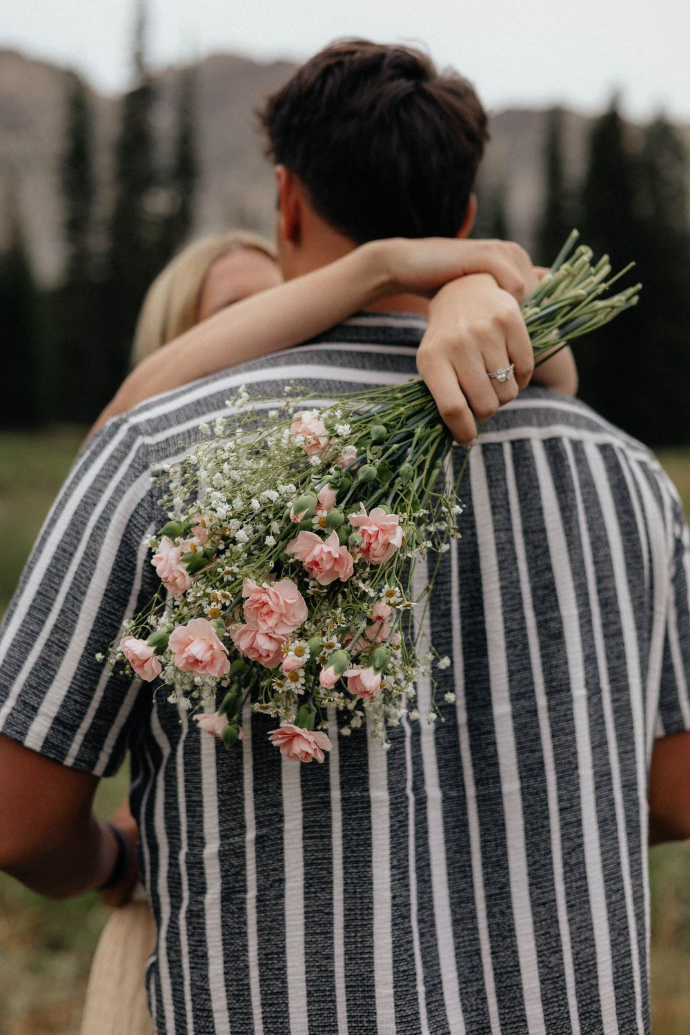 Couple embracing outdoors, woman holding a bouquet of pink roses and baby's breath, engagement ring visible.