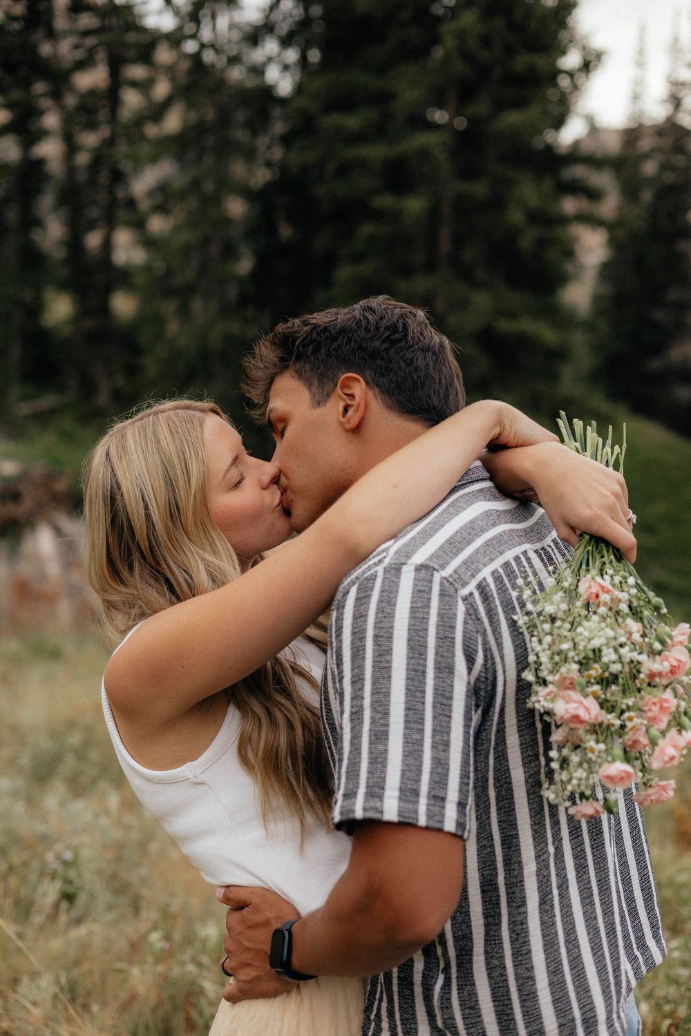 Couple sharing a romantic kiss outdoors, woman holding pink wildflowers, surrounded by tall evergreen trees.