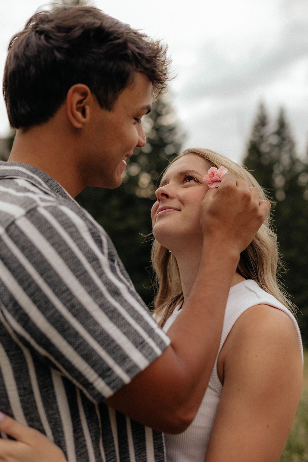 Man gently placing a pink flower in a smiling blonde womans hair in a scenic outdoor setting.