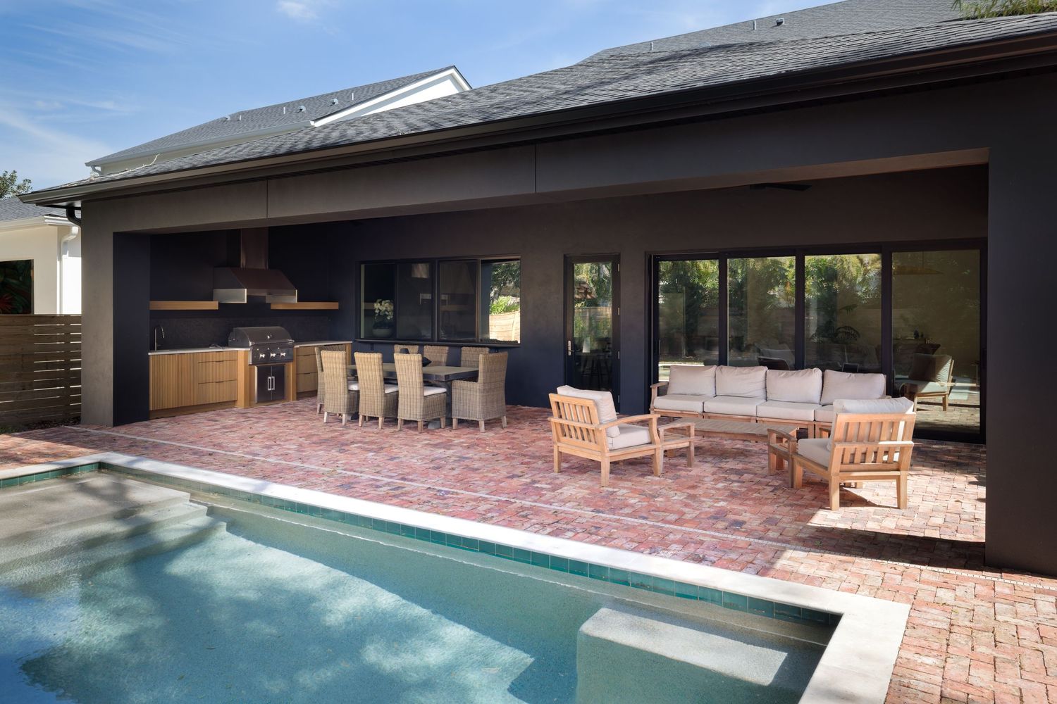 Modern dark outdoor living space with pool, brick patio, outdoor kitchen, and dining area.