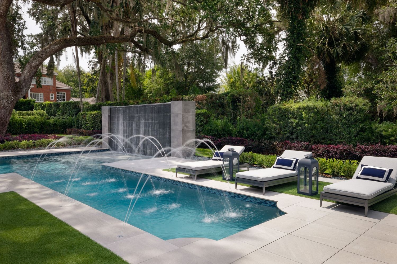 Luxury rectangular pool with fountain jets, lounge chairs, lush tropical landscaping, and a water wall feature.