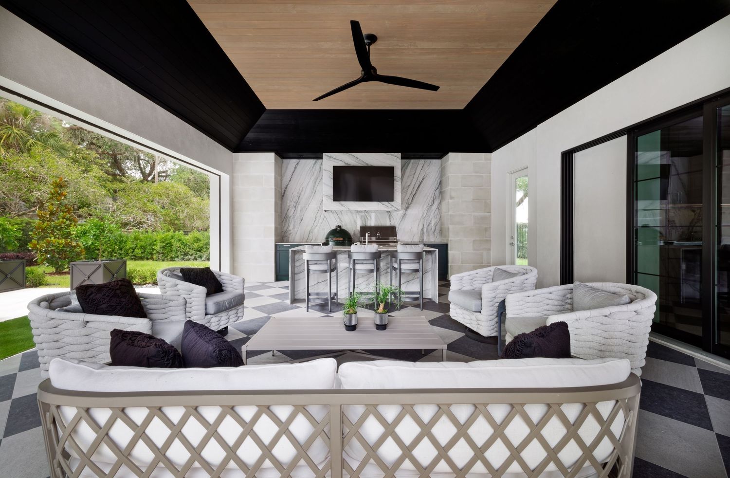 Sophisticated covered outdoor lounge with marble fireplace, checkered floor, white sofas, and ceiling fan.