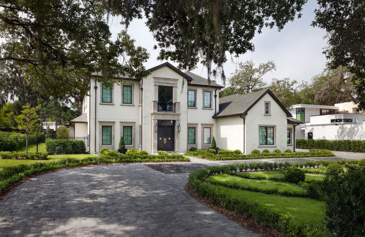 Elegant white estate with teal shutters, circular driveway, manicured hedges, and towering oak trees.