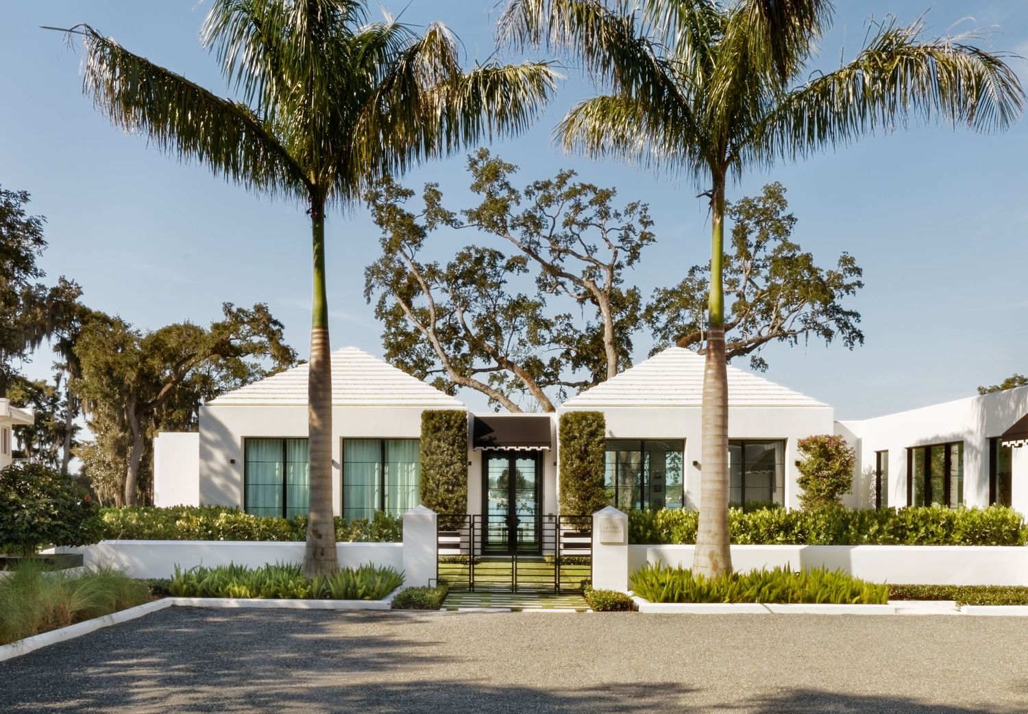 Modern white luxury home with palm trees, manicured hedges, and glass double doors at golden hour.