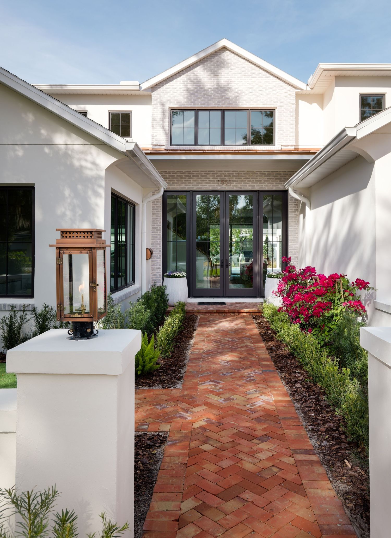 Charming white brick home entrance with herringbone brick pathway, black French doors, and blooming flowers.