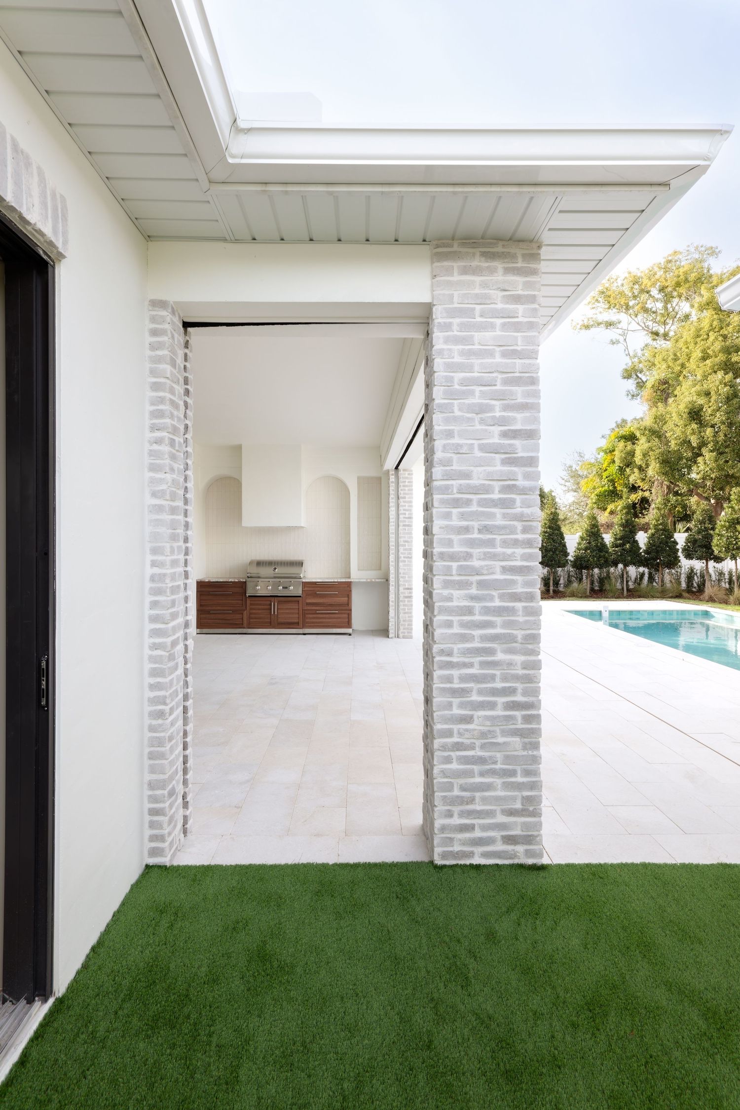 Modern white brick outdoor covered kitchen area with wood cabinetry, green lawn, and pool view.