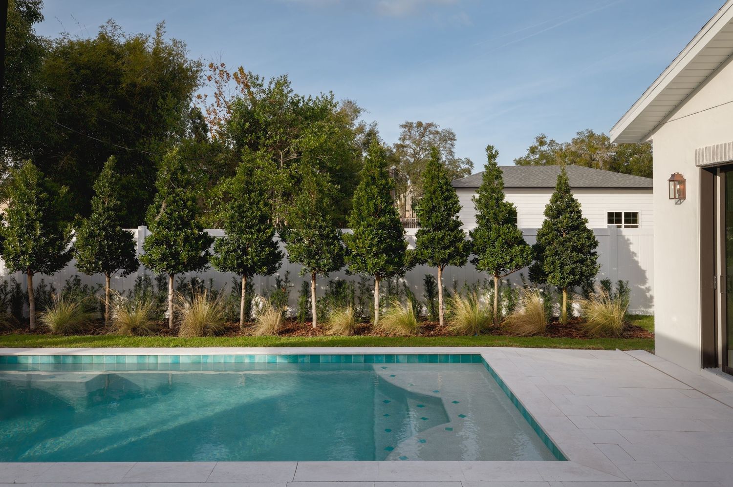 Clean modern rectangular swimming pool surrounded by tall columnar trees and white fence landscaping.