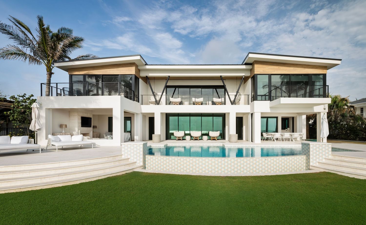 Luxury white modern beachfront mansion with large swimming pool, manicured lawn, and expansive glass windows.