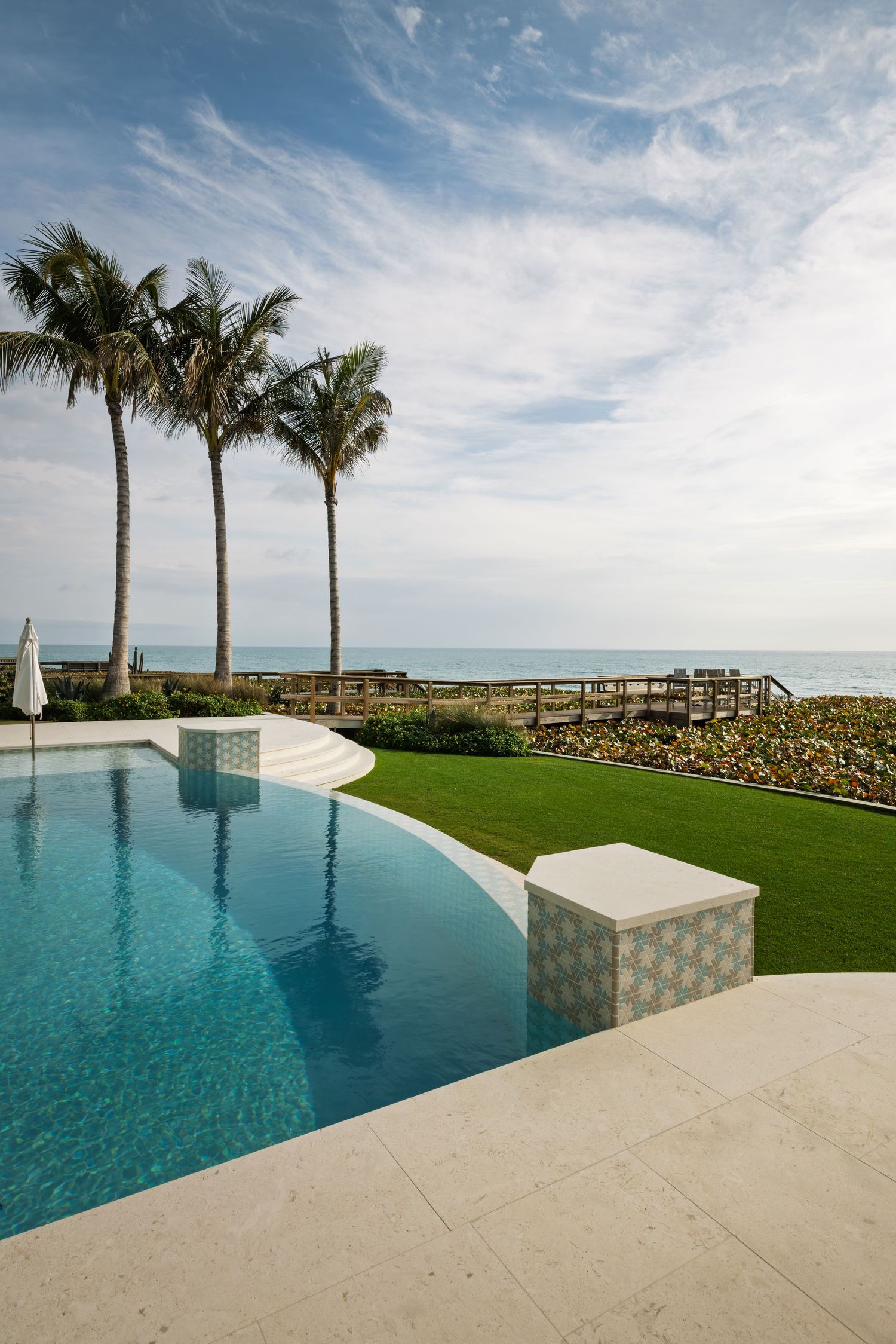 Elegant infinity pool with palm trees and ocean view under blue sky at luxury beachfront estate.