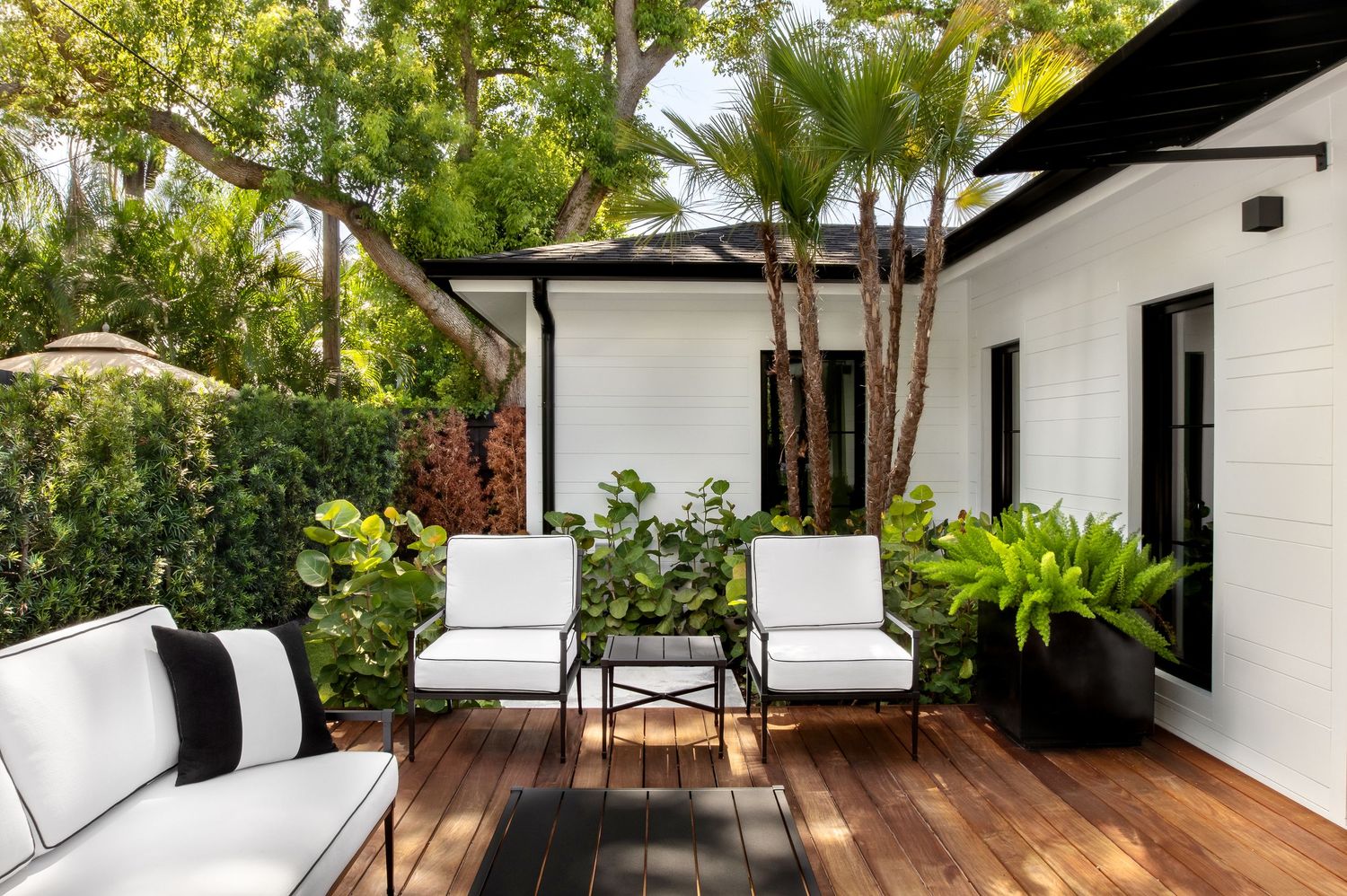 Charming white cottage patio with wood deck, white lounge furniture, lush tropical plants, and palm trees.