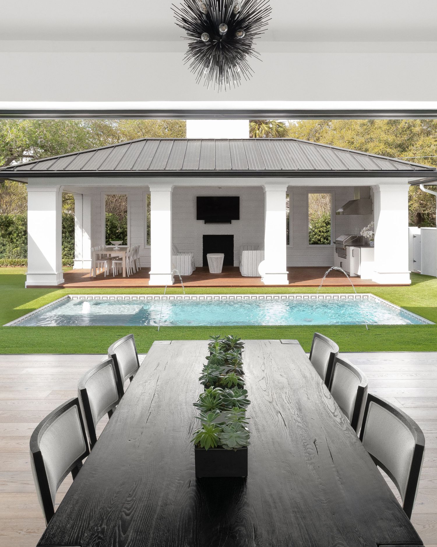 Luxury outdoor dining area with black table overlooking a rectangular pool and white poolhouse with columns.