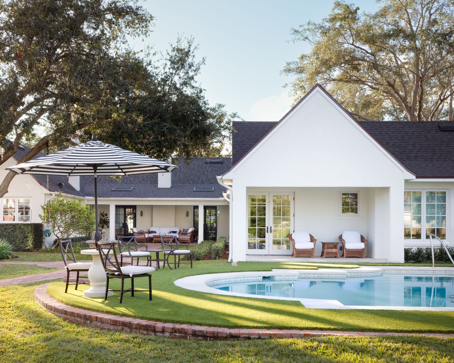 White cottage-style home with oval pool, covered patio seating, and outdoor dining area on lush green lawn.
