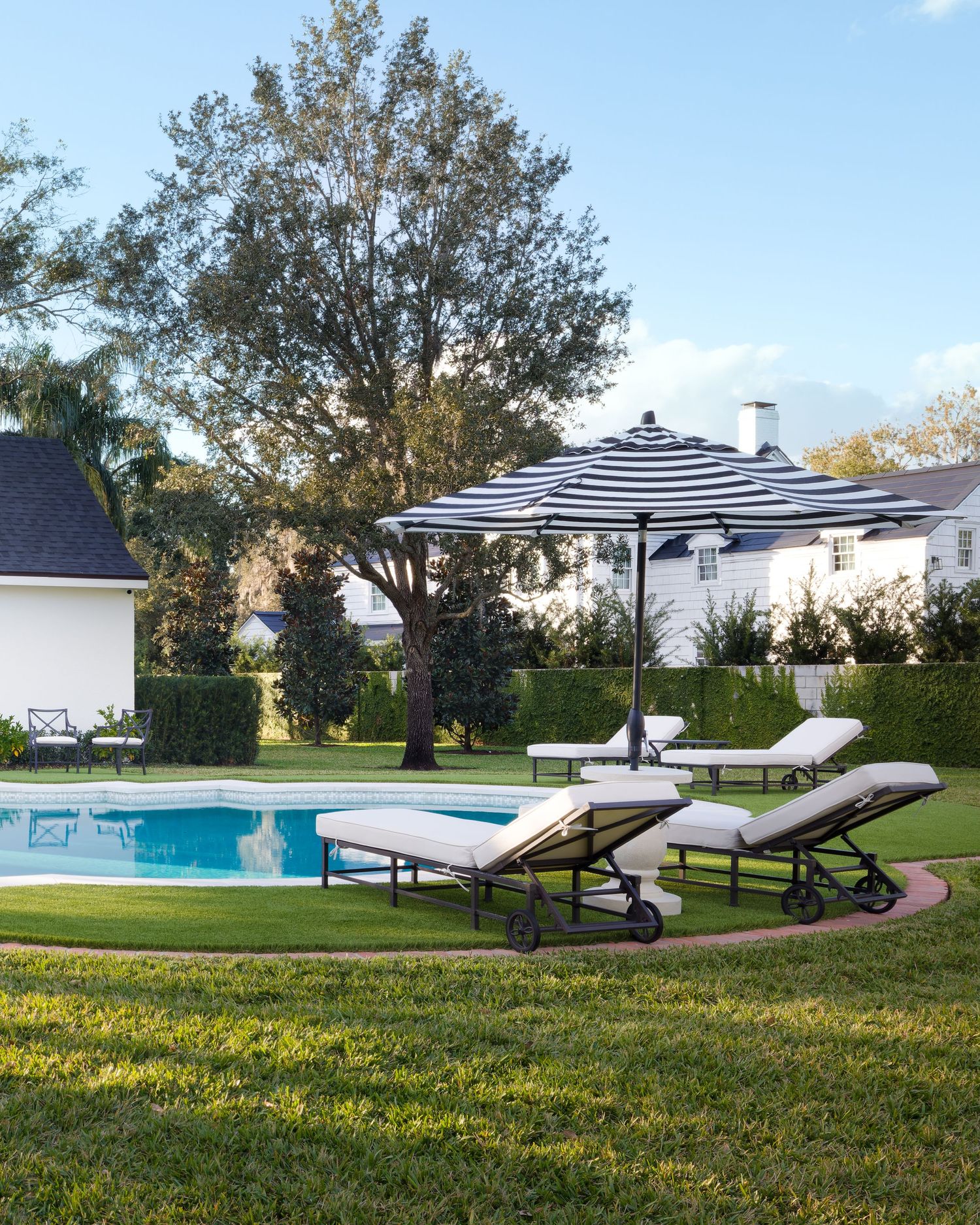 Elegant backyard pool with navy striped umbrella, lounge chairs, and white home surrounded by mature trees.