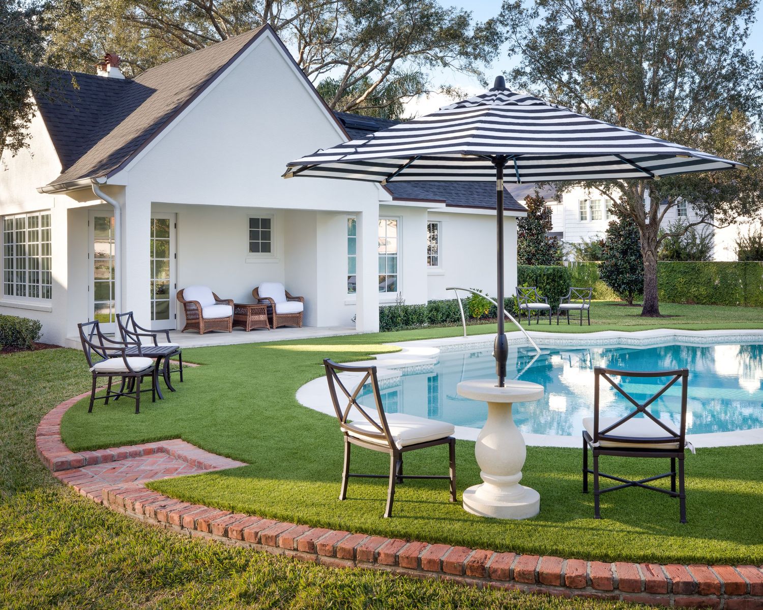 Bright backyard pool area with navy striped umbrella, bistro chairs, white pedestal table, and manicured lawn.