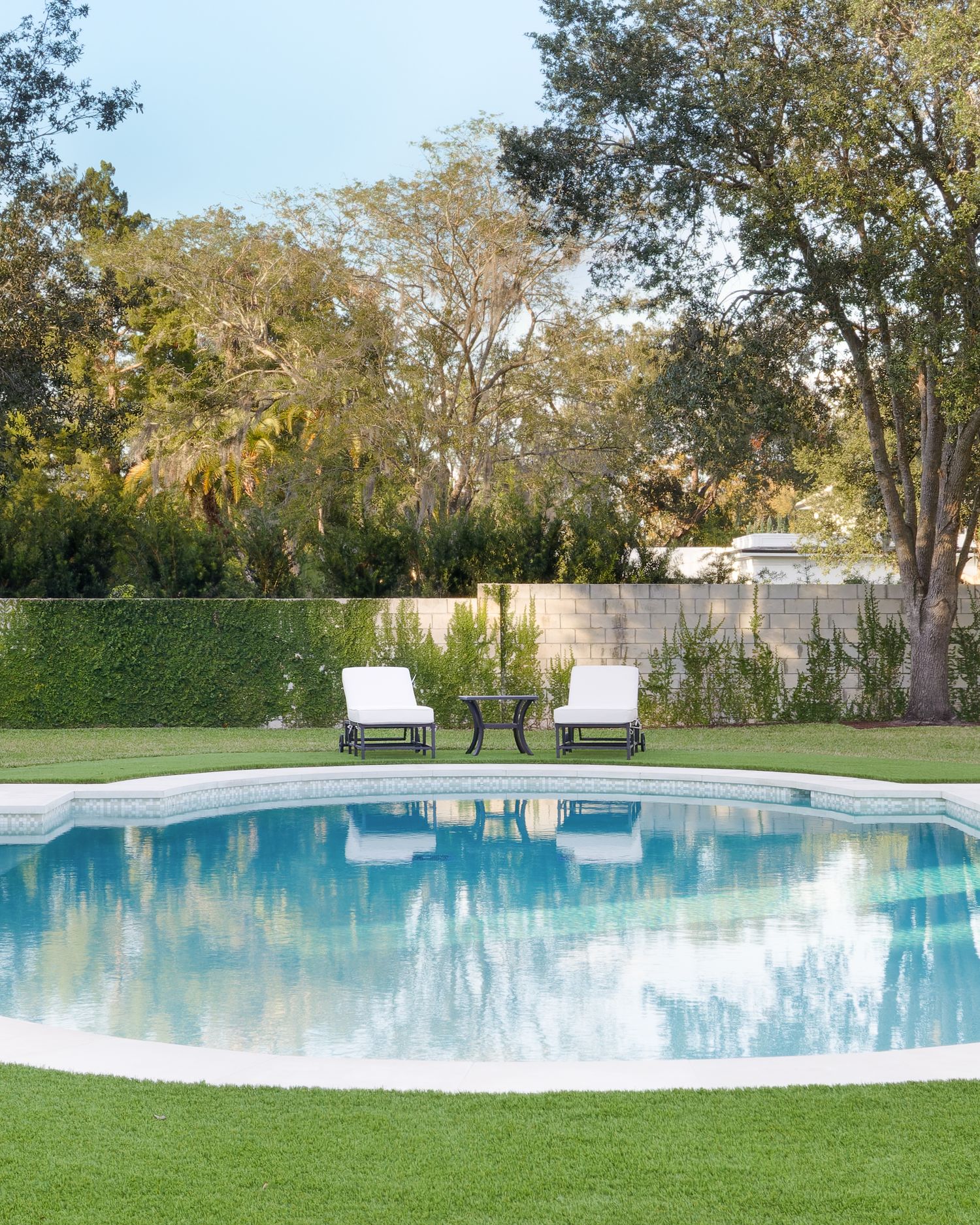 Serene oval swimming pool with white lounge chairs reflecting tall trees in calm blue water on green lawn.