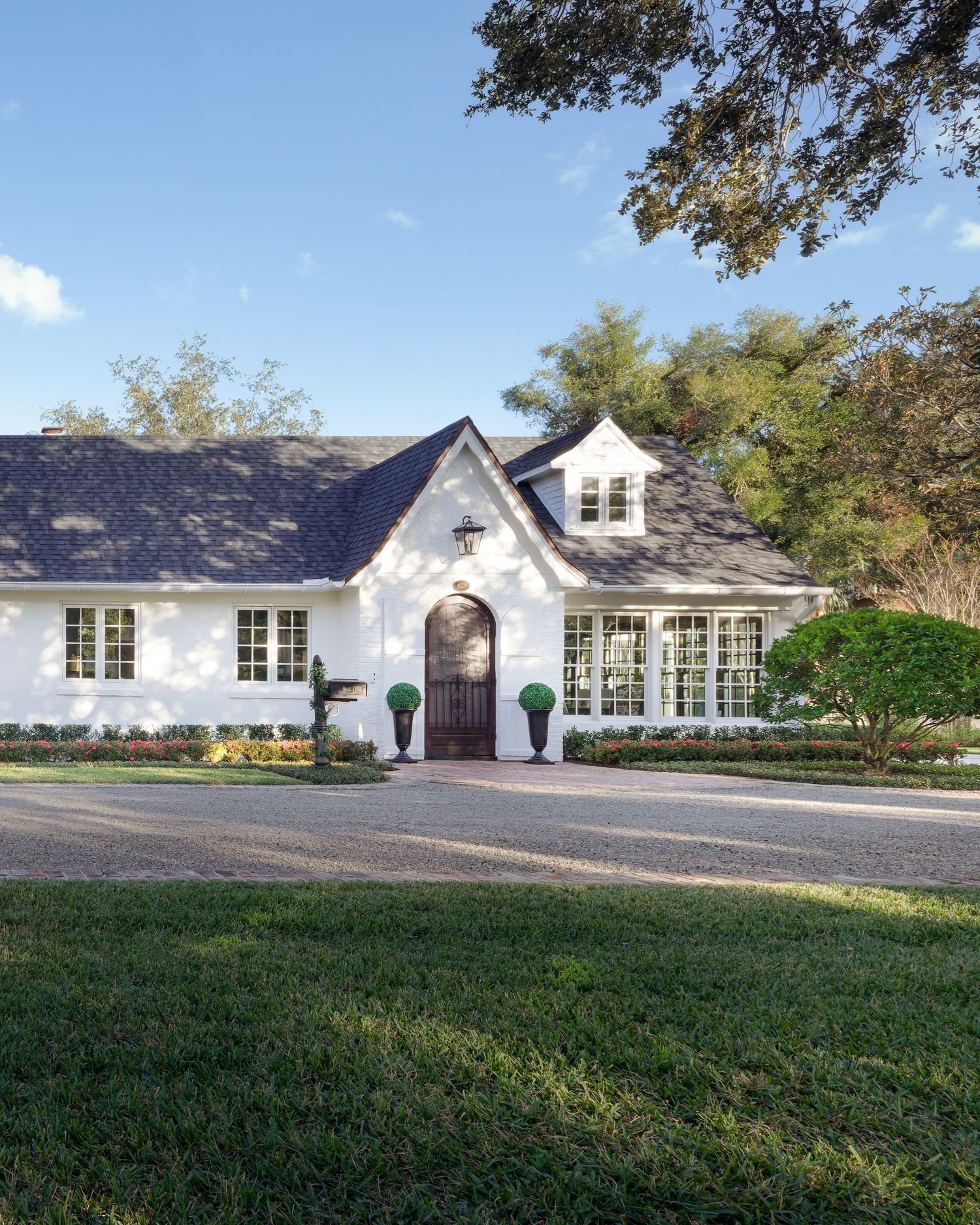 White French country home with arched wood door, manicured topiaries, and dark shingled roof surrounded by lush trees.