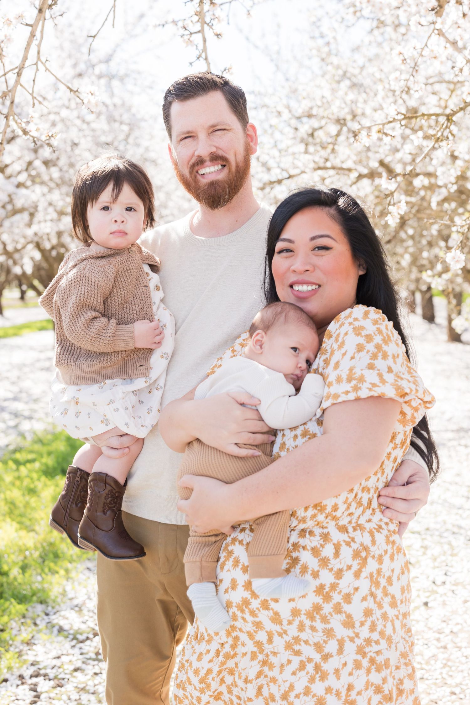 Happy family of four posing outdoors among blooming white trees, parents holding two young children in spring setting.