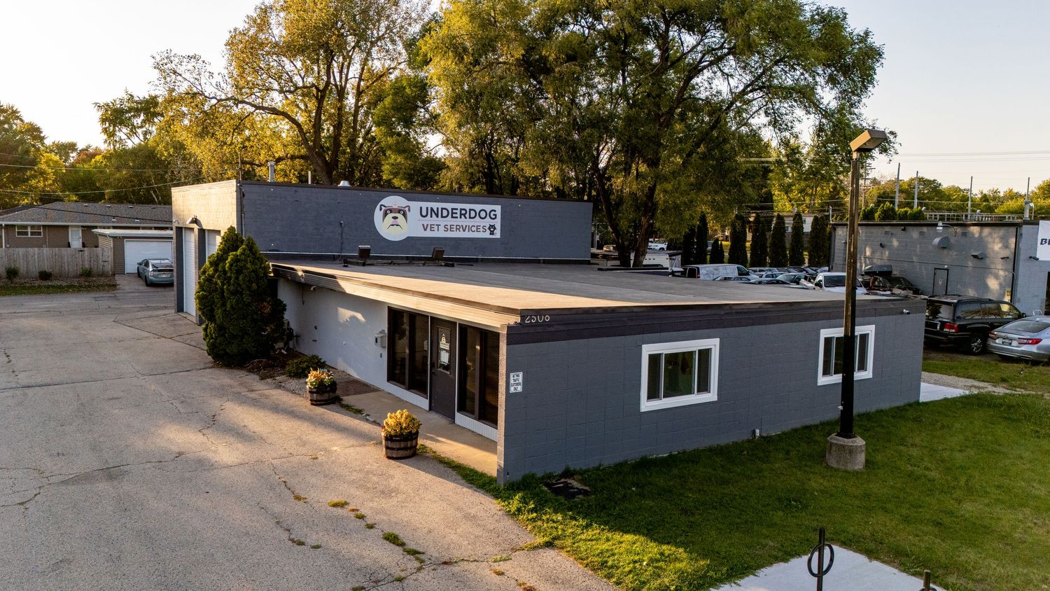 Aerial view of Underdog Automotive shop, a modern gray commercial building with signage, parking lot, and trees at sunset.