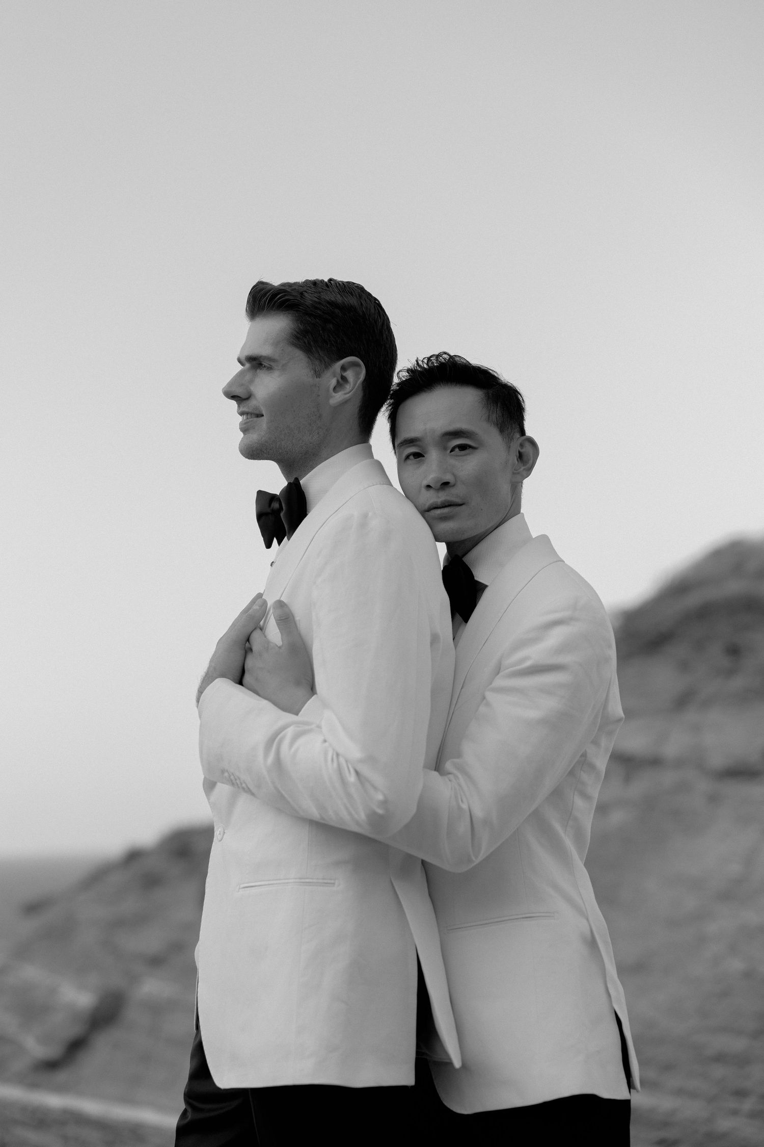 Same sex couple destination wedding LGBTQ+ photographer white tuxedo jackets embrace outdoors romantic black and white wedding portrait.