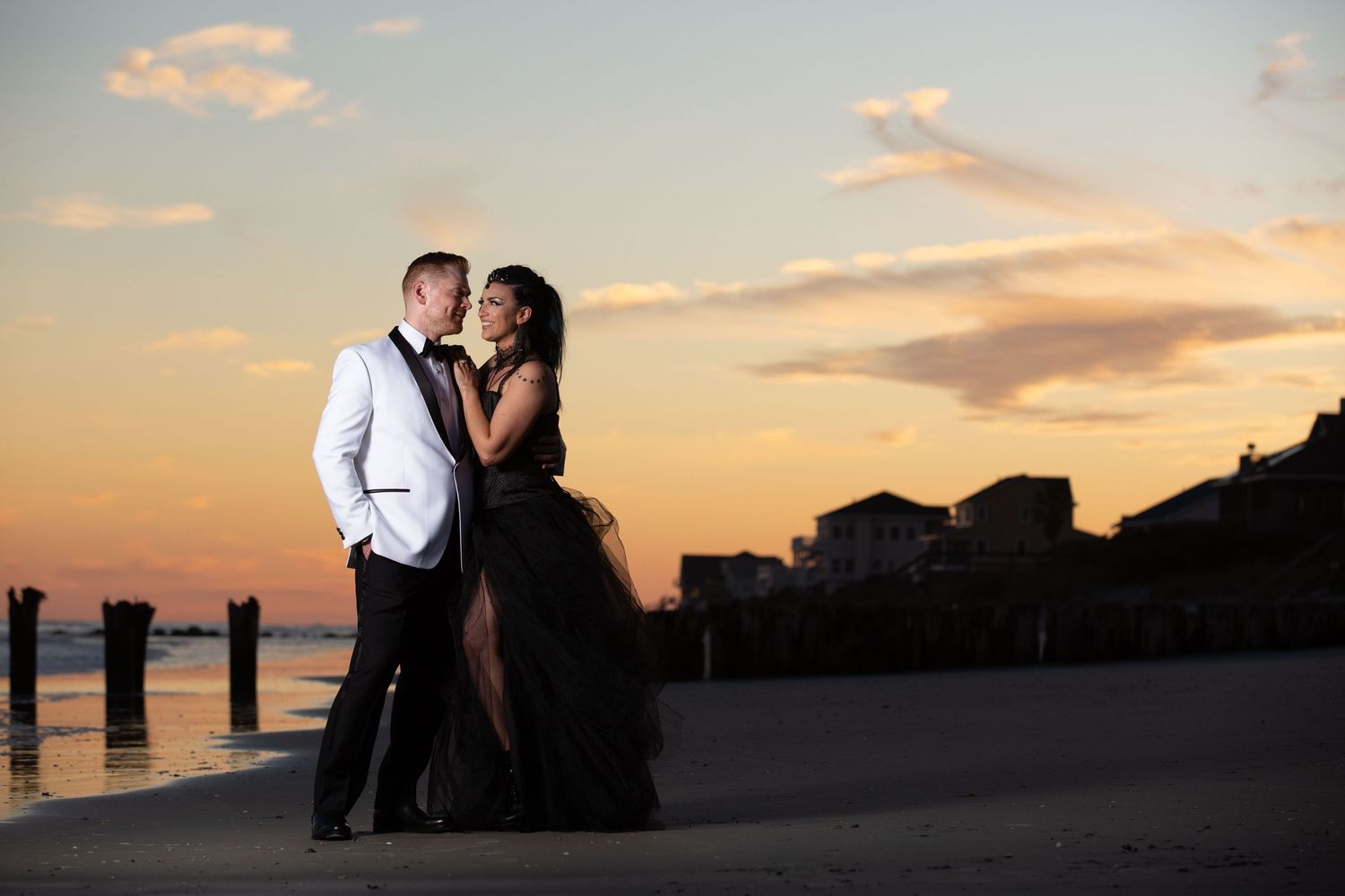 Couple in formal wear embracing on beach at sunset, man in white tuxedo, woman in black gown.