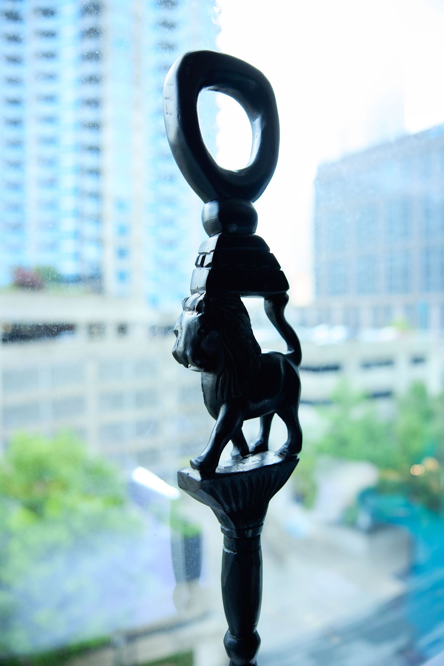 Dark carved wooden African figurine featuring a rider on horseback with loop top, displayed against urban cityscape backdrop.