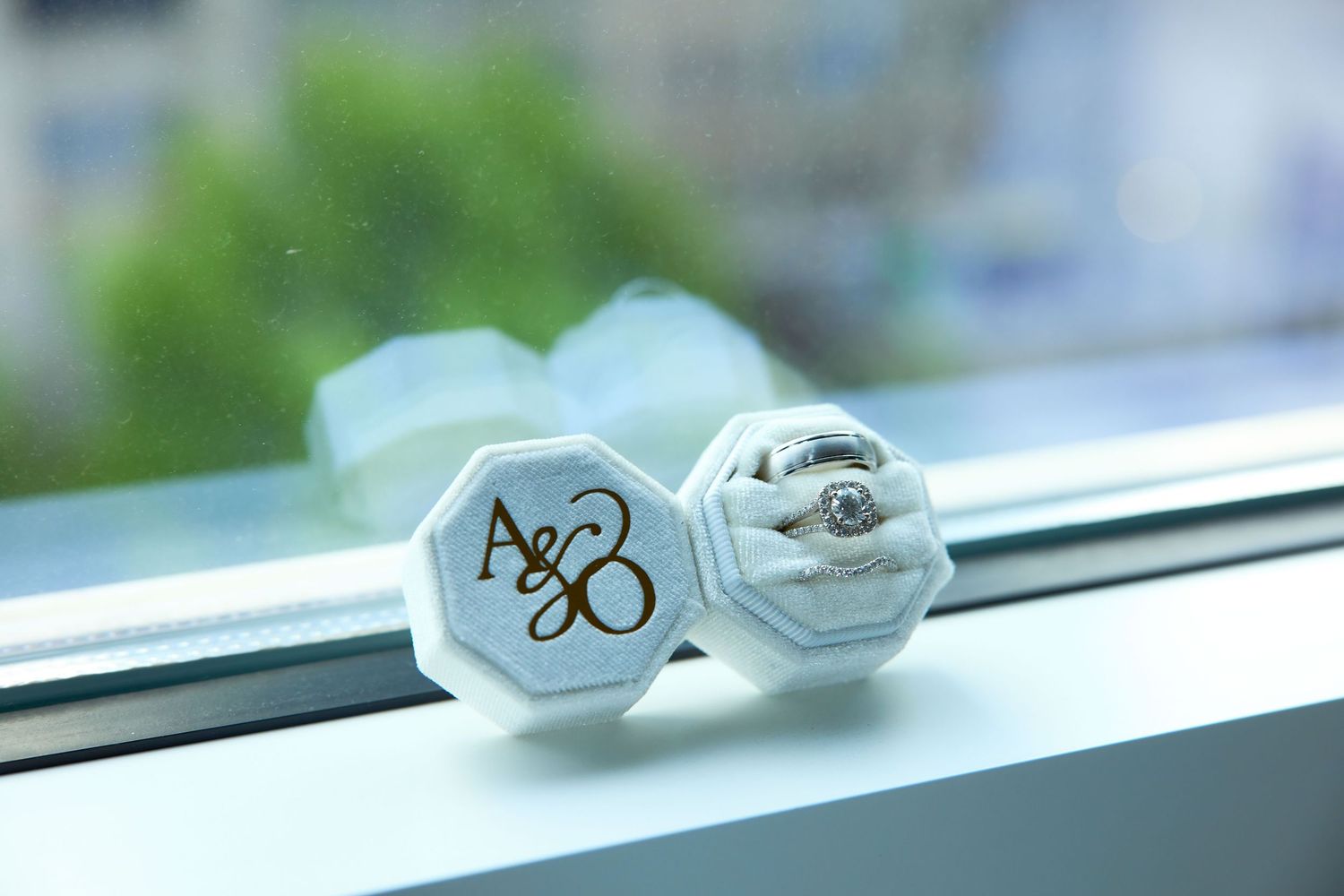 White octagonal ring box monogrammed with A&B initials, holding a diamond engagement ring on a windowsill.