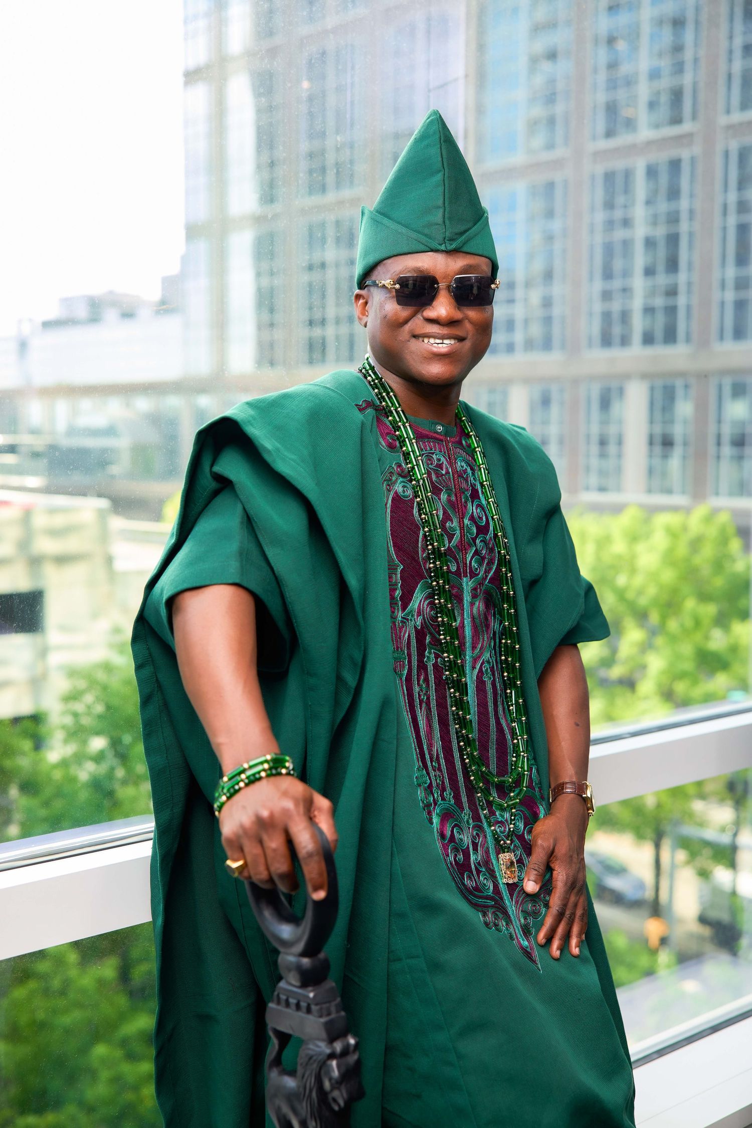 Man in green traditional African agbada robe and matching fila hat smiling outdoors near modern building.