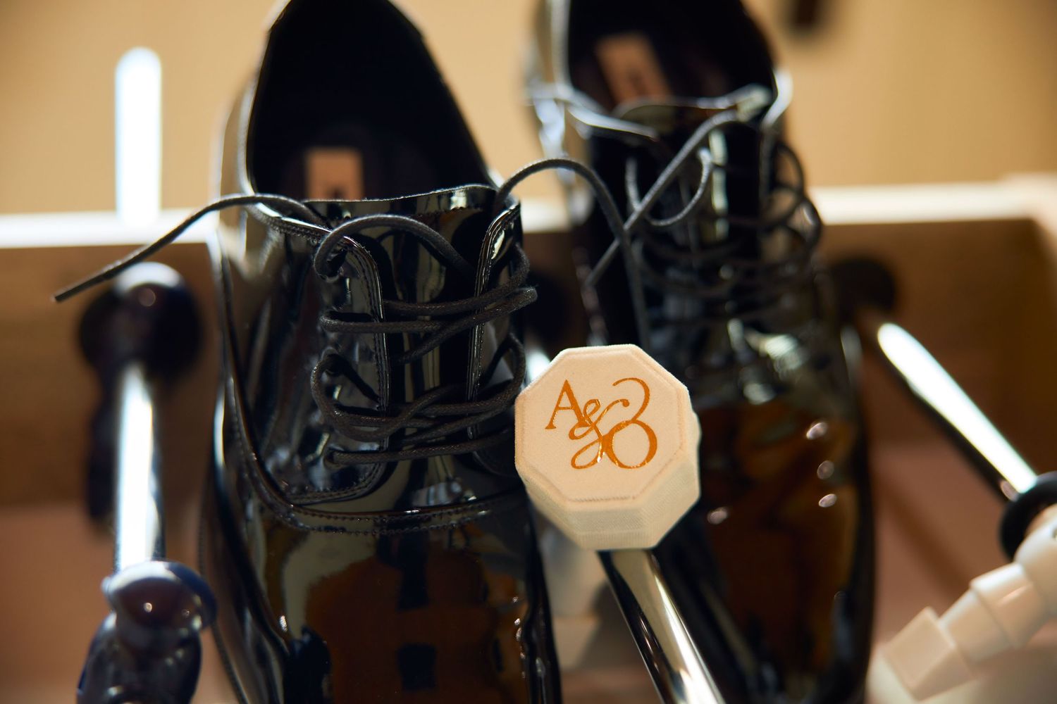 Shiny black dress shoes with white laces beside a monogrammed octagonal wedding detail on a wooden rack.