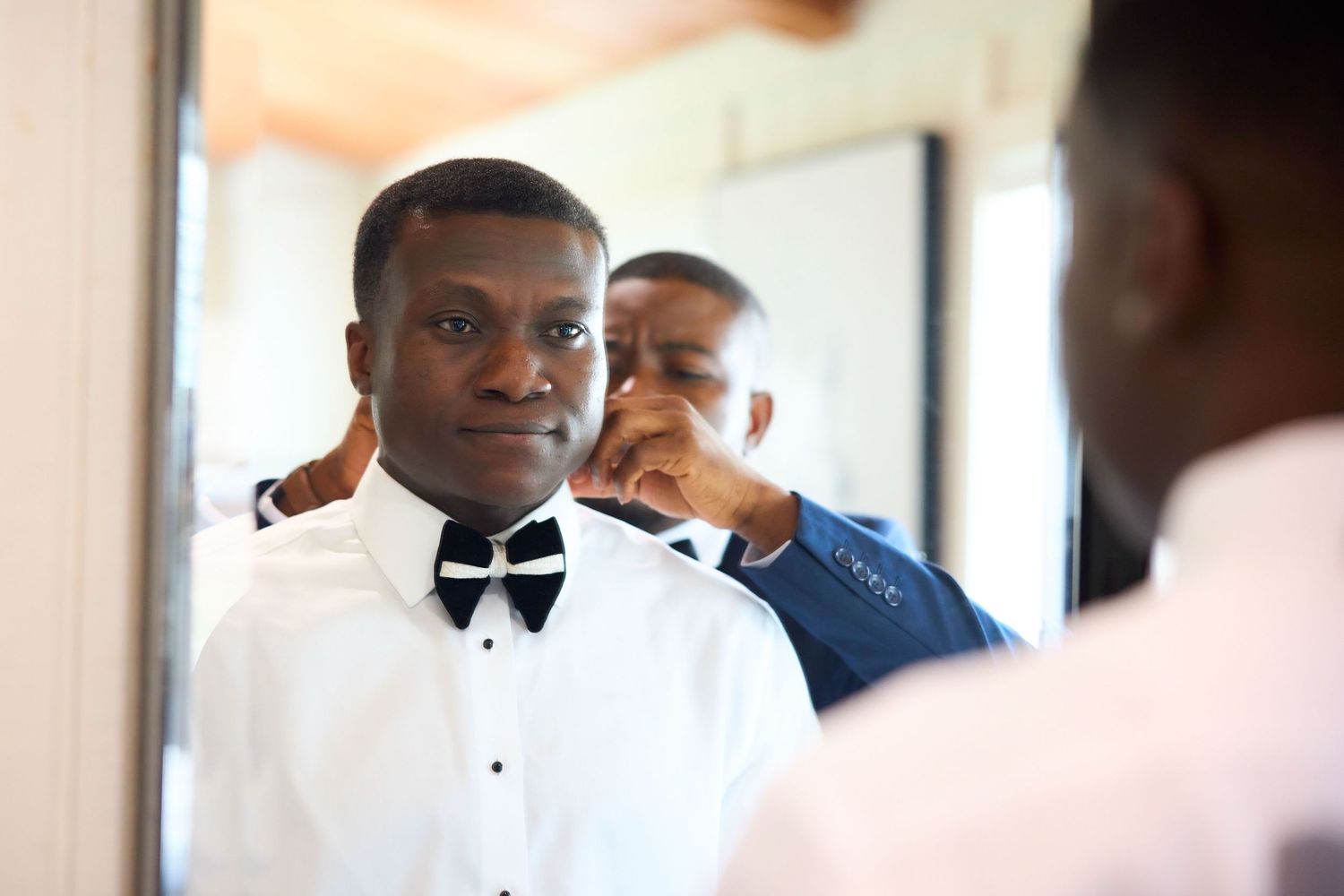A man in a white tuxedo shirt and black bow tie gets dressed with help from another man in a mirror.