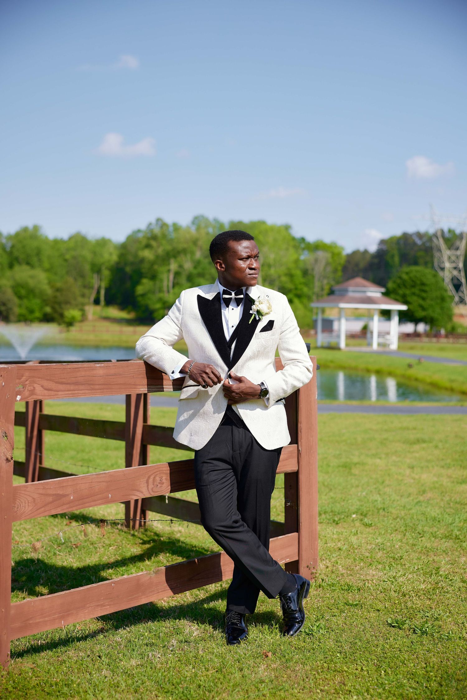 Elegant Black man in white tuxedo jacket and bow tie leans against wooden fence at outdoor wedding venue.