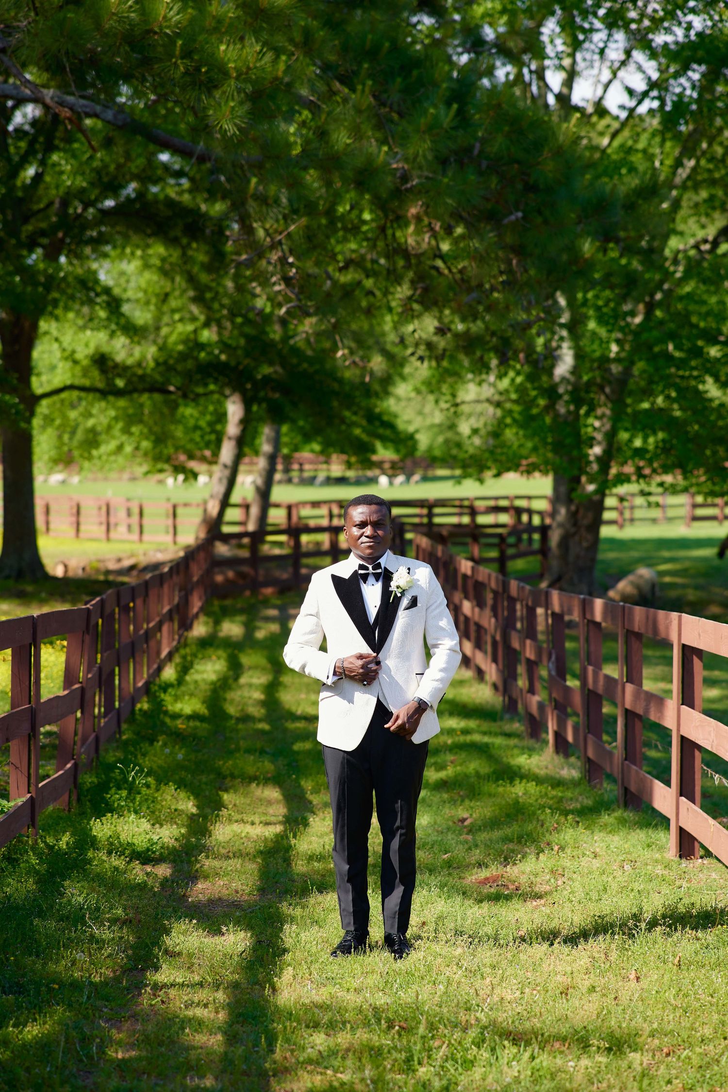A groom in a white tuxedo jacket walks between wooden fences on a lush green farm path on his wedding day.