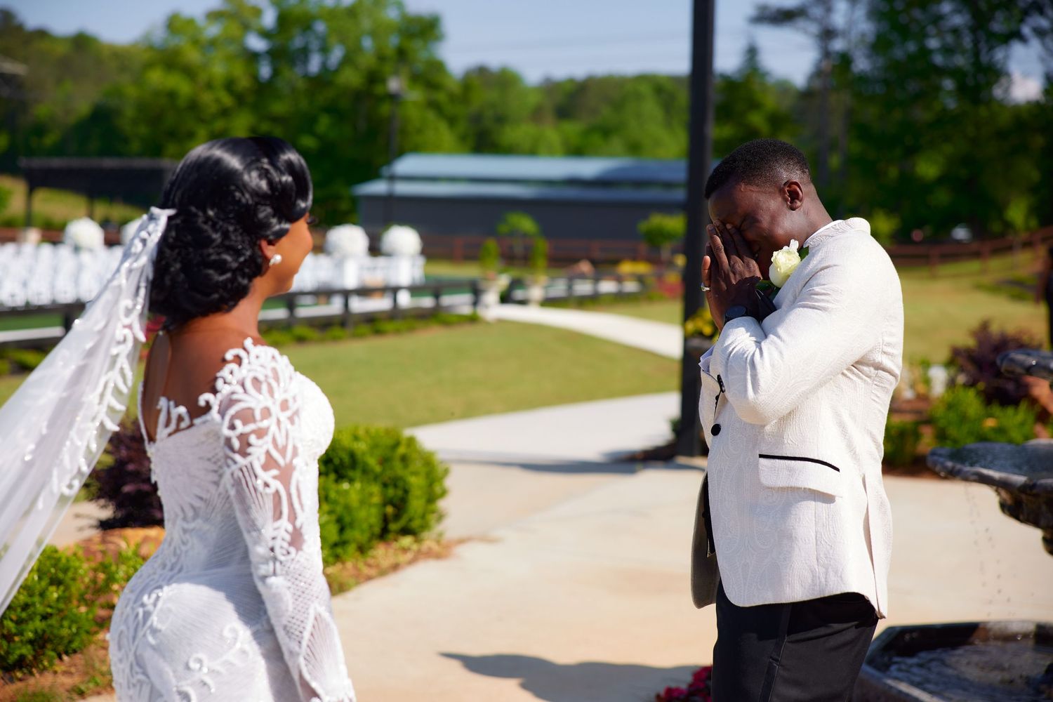 Emotional groom in white tuxedo wipes tears seeing bride in lace wedding dress during outdoor first look ceremony.