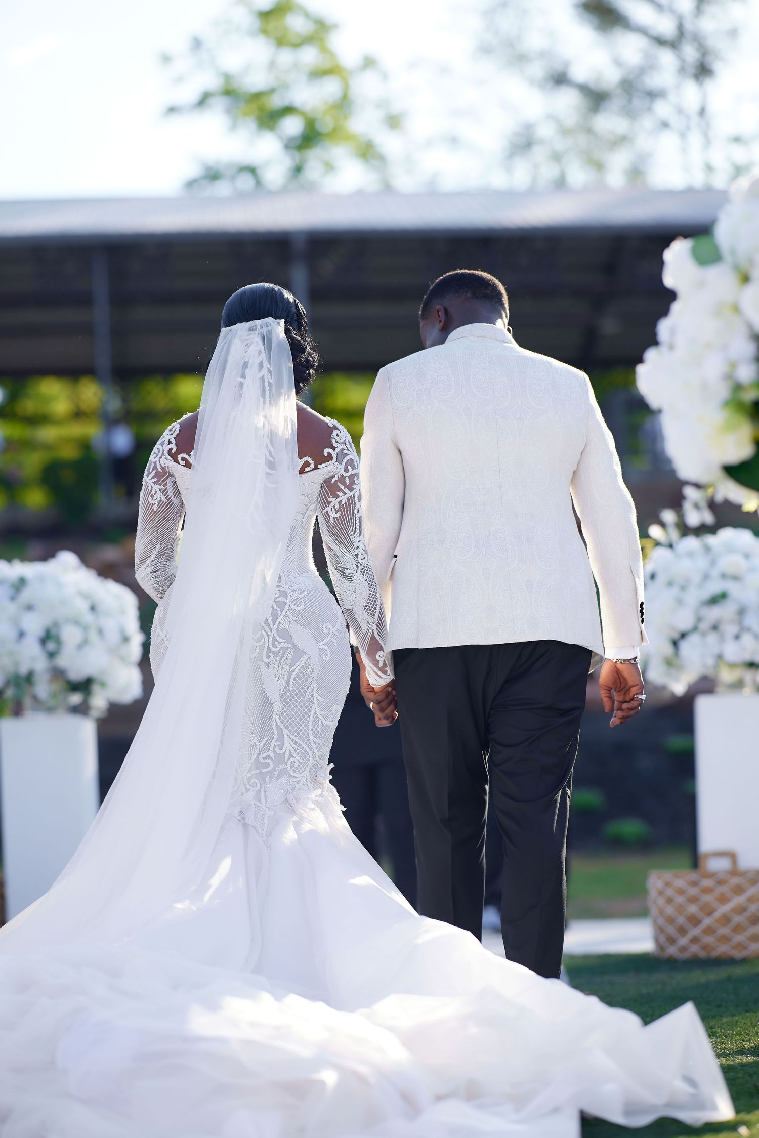 Bride and groom holding hands at outdoor wedding ceremony, bride in lace gown with veil, groom in white jacket.