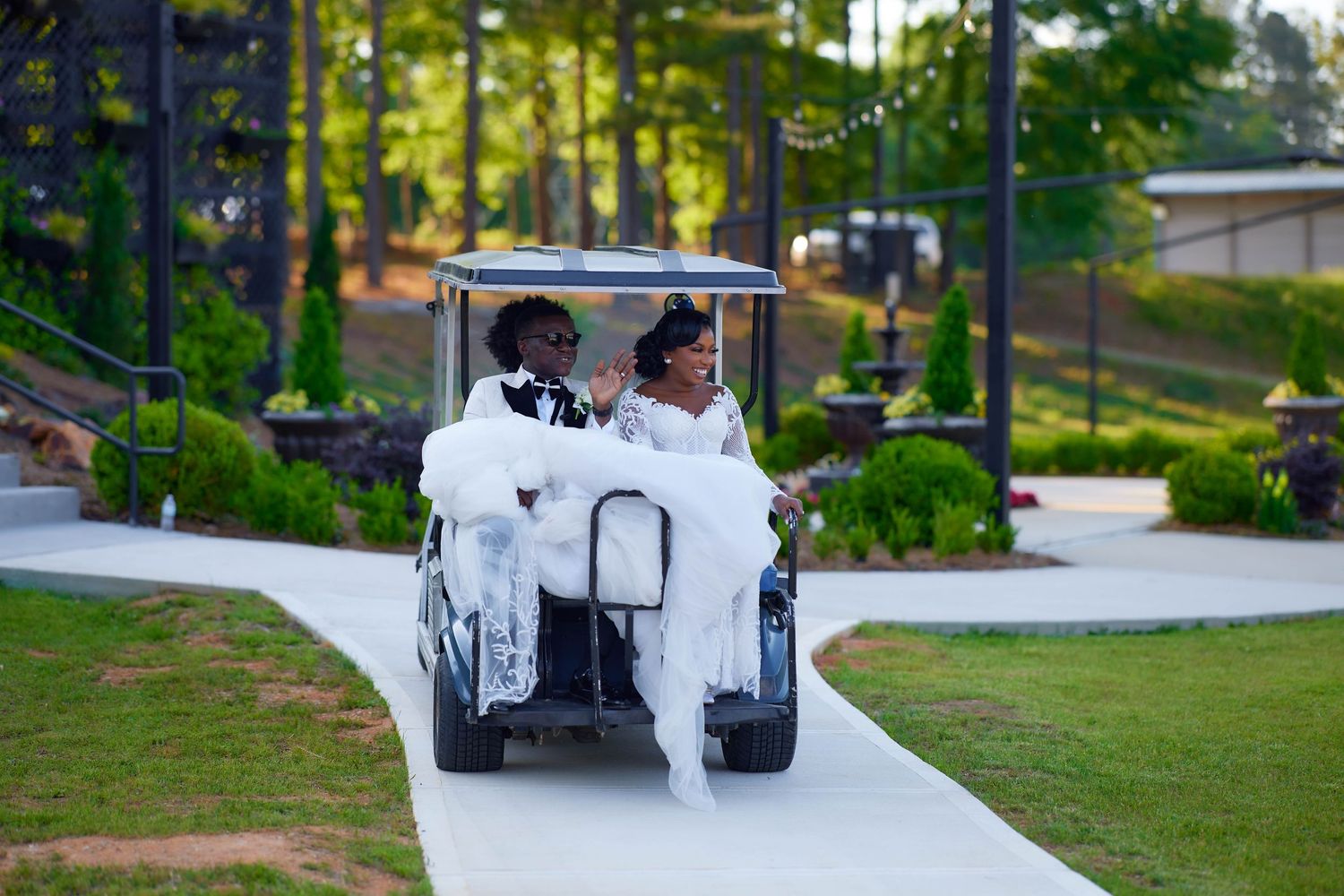 Bride and groom riding a decorated golf cart down a white path at an outdoor wedding venue.