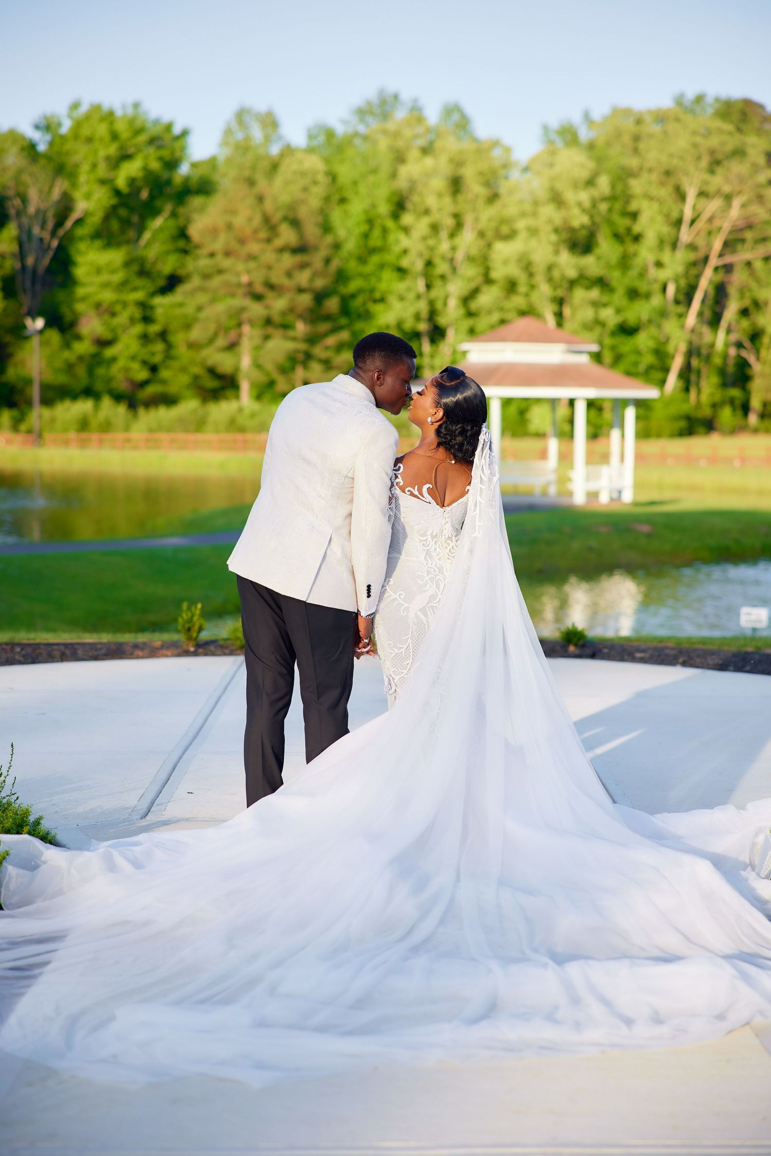 Newlywed couple sharing a kiss in wedding attire by a serene pond with a gazebo and lush green trees backdrop.