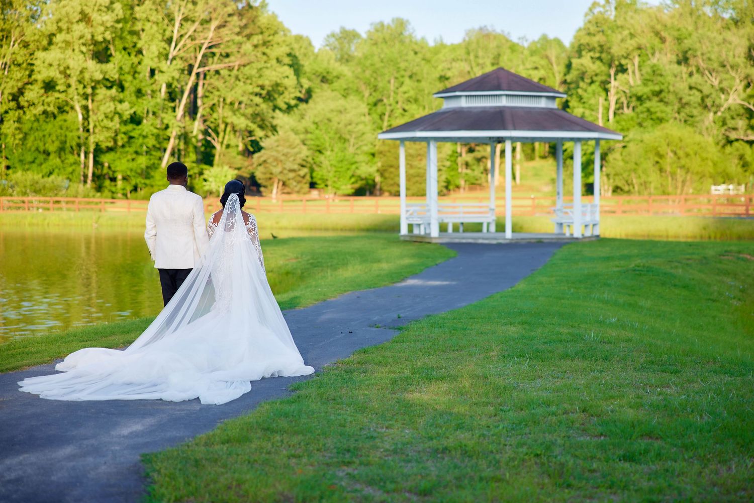 Bride and groom walking toward a white gazebo by a serene pond surrounded by lush green trees on their wedding day.