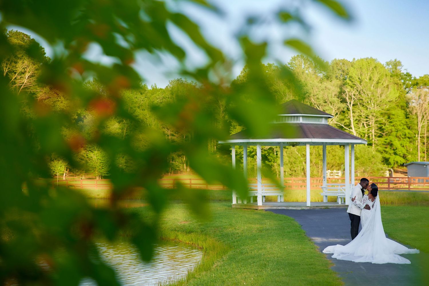 Bride and groom embracing near a white gazebo by a pond surrounded by lush green trees on a sunny wedding day.