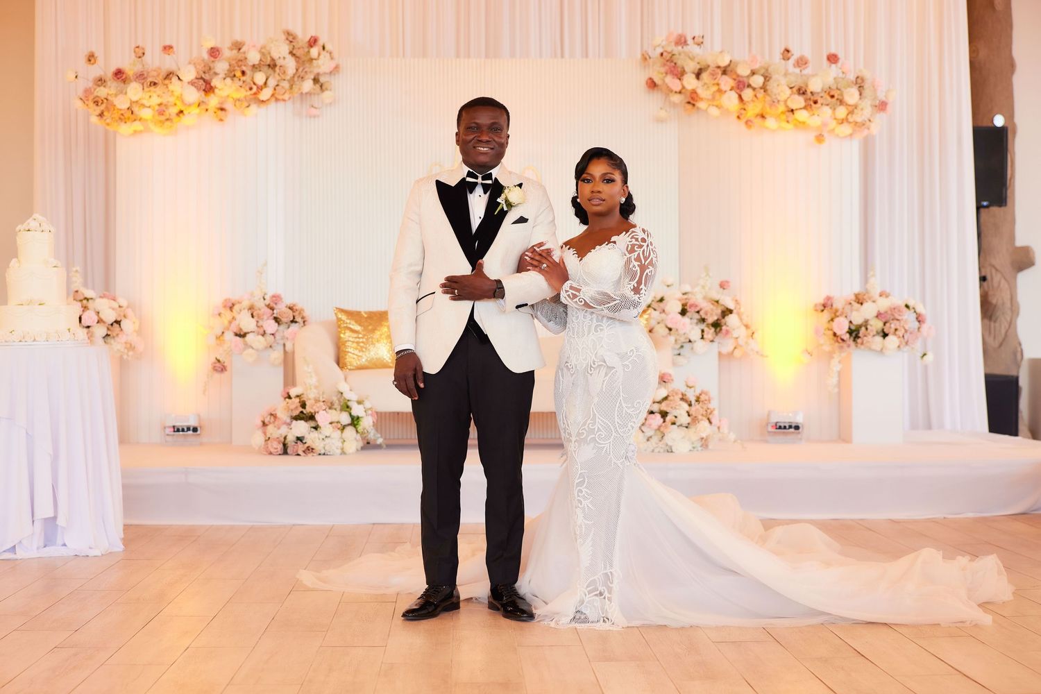 Elegant wedding couple posing at reception venue with floral backdrop, white gown, and tuxedo.