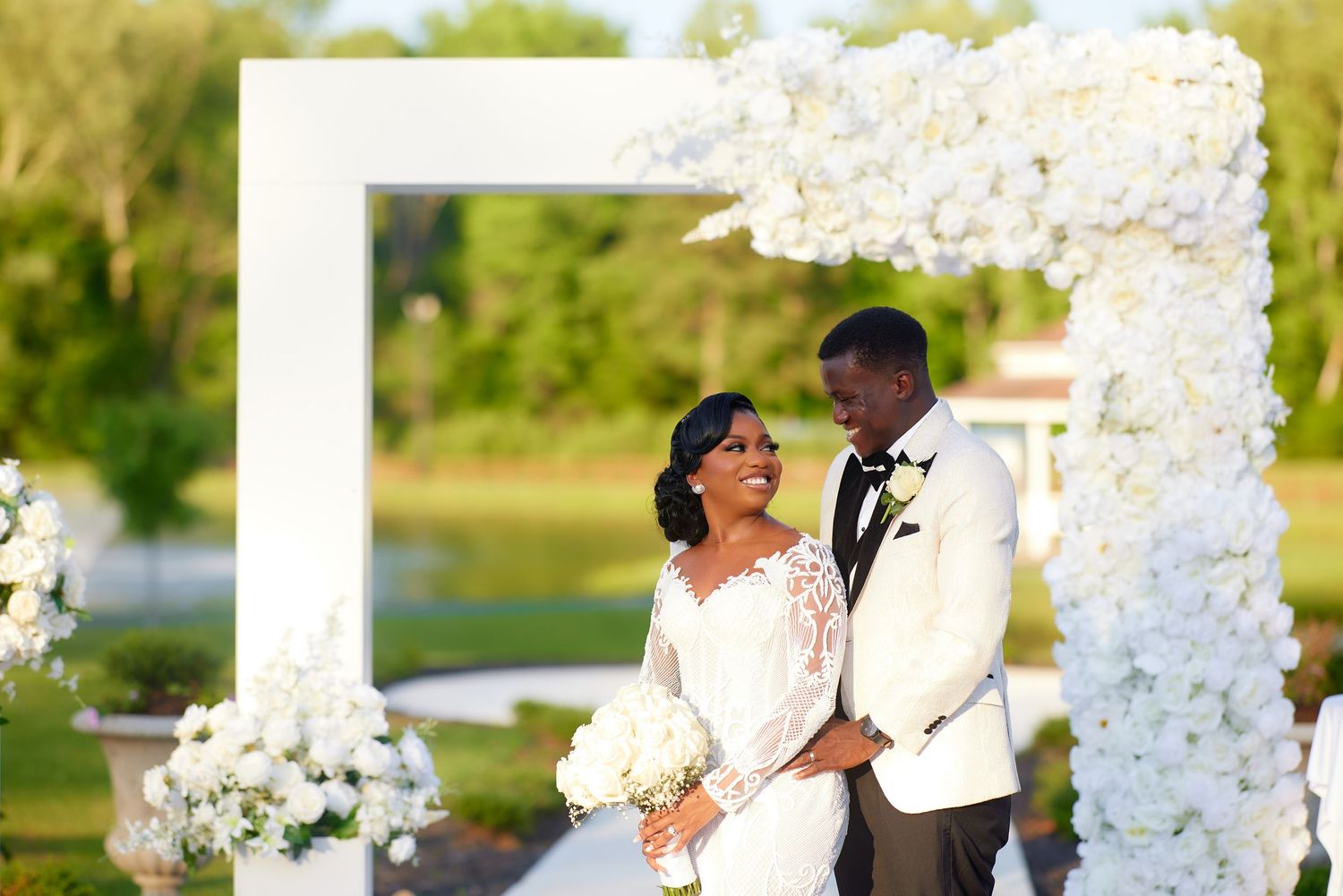 A couple poses under a white floral arch at an outdoor wedding ceremony, bride holding a bouquet.