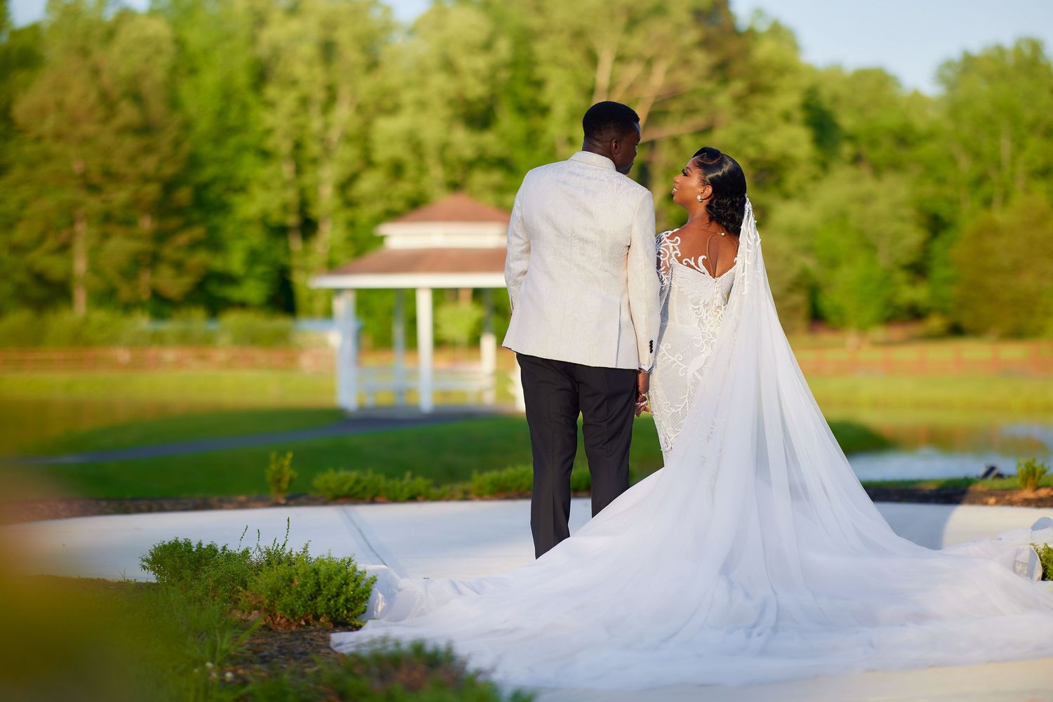 Bride and groom holding hands facing a scenic garden gazebo, bride wearing elegant lace wedding gown with long veil.