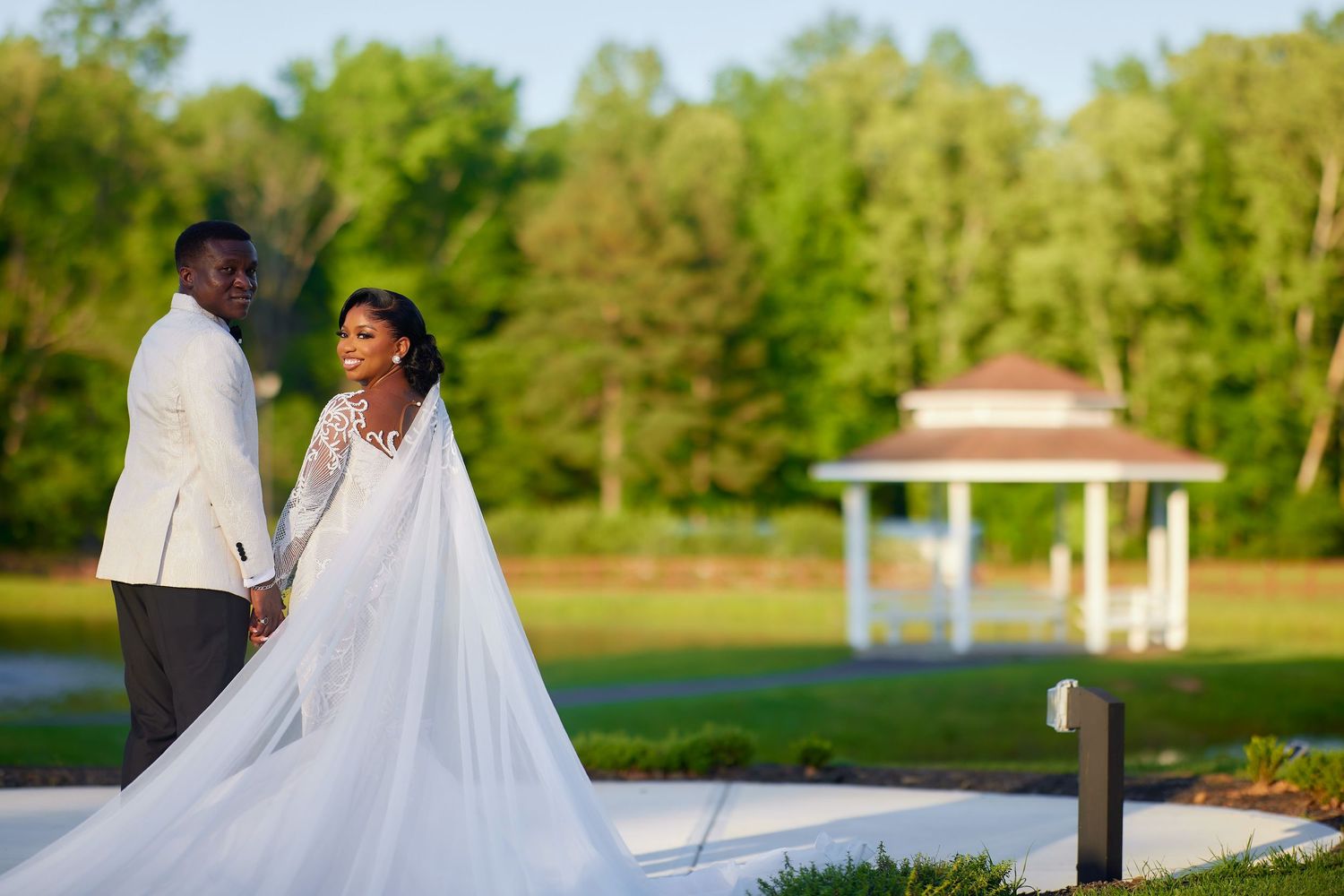 Elegant bride and groom holding hands outdoors, bride in long white gown with train, groom in white suit, gazebo background.