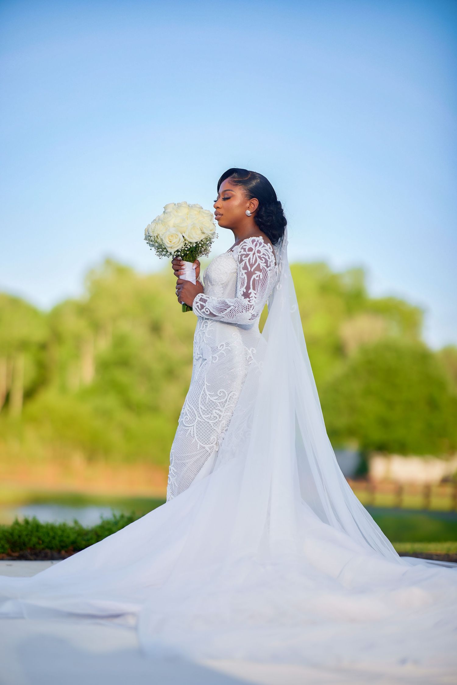 Elegant bride in a lace long-sleeve mermaid gown with cathedral veil holding white bouquet outdoors.
