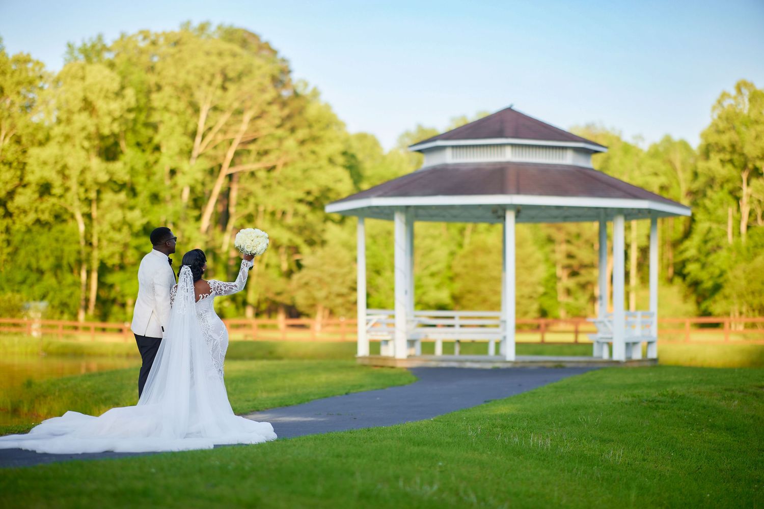 Bride and groom stand near a white gazebo on a lush green lawn during a sunny outdoor wedding ceremony.