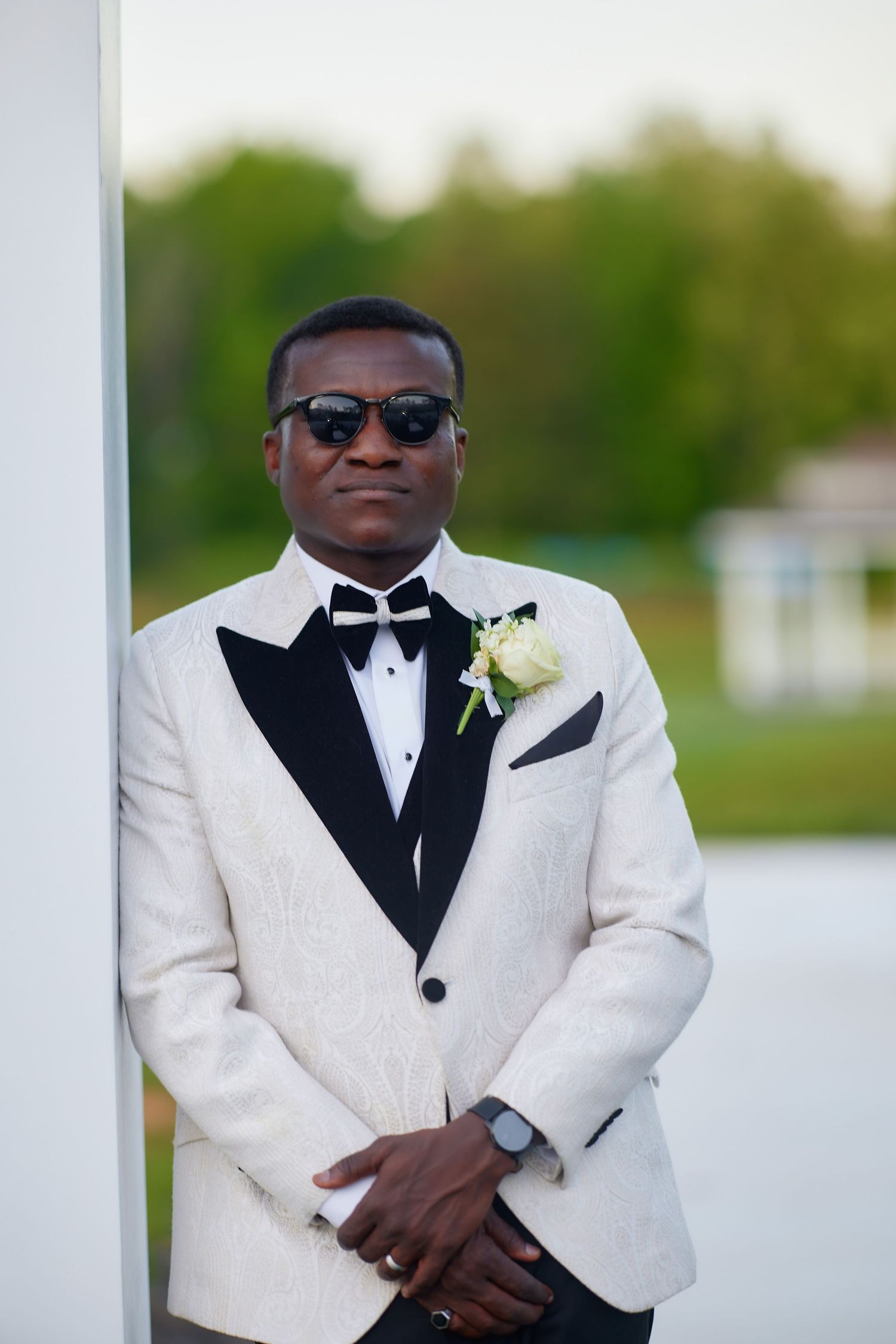 Elegant groom in white tuxedo jacket with black bow tie and boutonniere, wearing sunglasses outdoors.