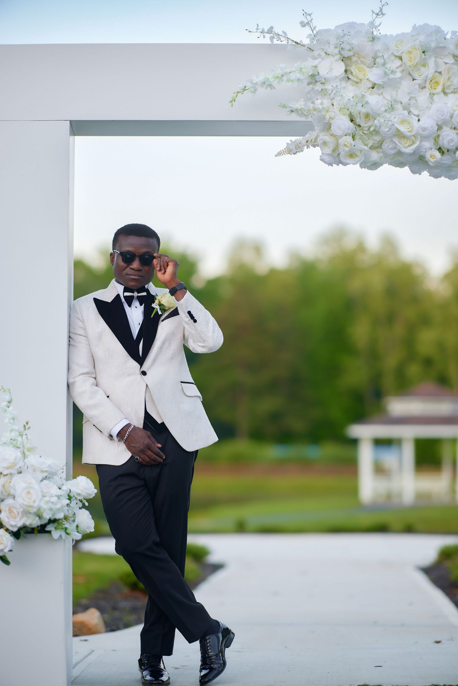 Elegant groom in white tuxedo jacket leaning against floral wedding arch outdoors.
