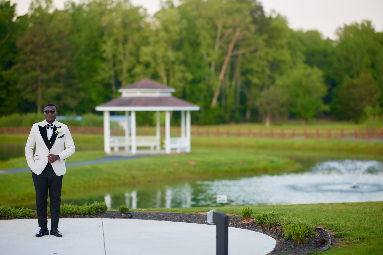 Groom in white tuxedo jacket stands on curved path near pond and white gazebo surrounded by lush green trees.