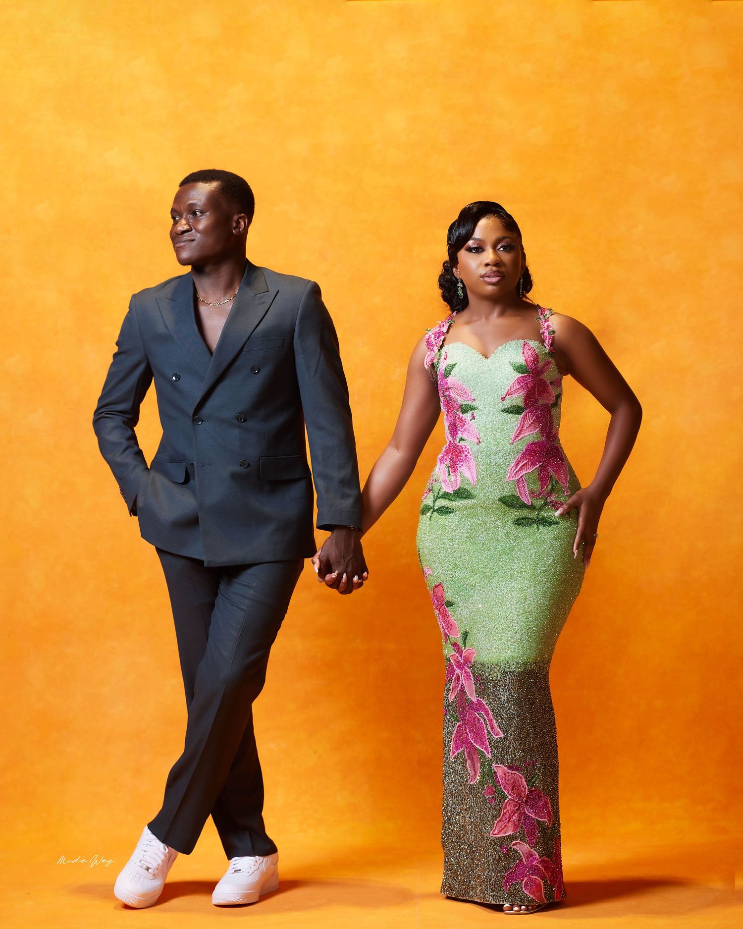 Couple posing together, man in dark suit holding hands with woman in green and pink floral African print gown, orange background.