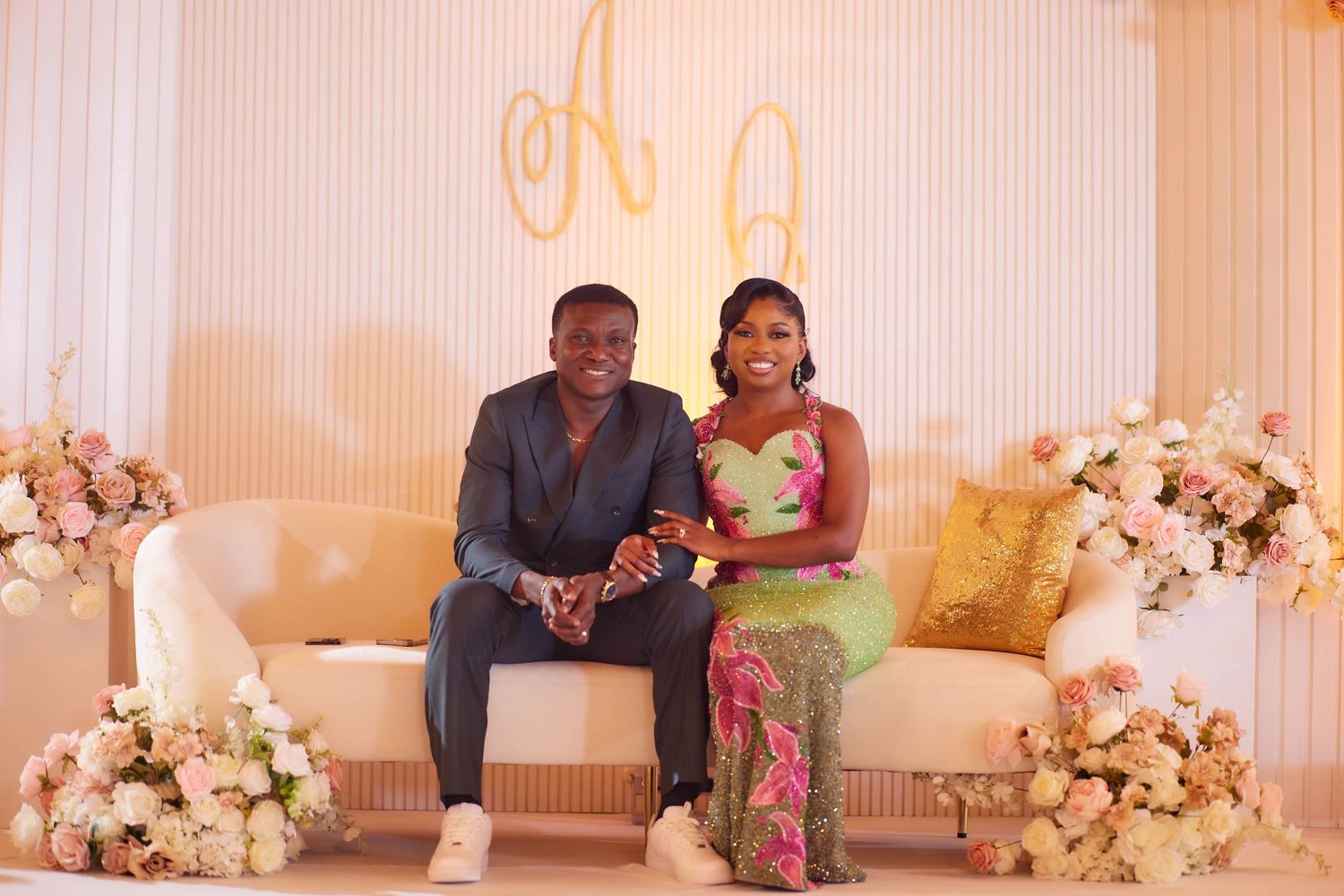 Couple in elegant attire seated on white sofa with floral arrangements and gold monogram backdrop.