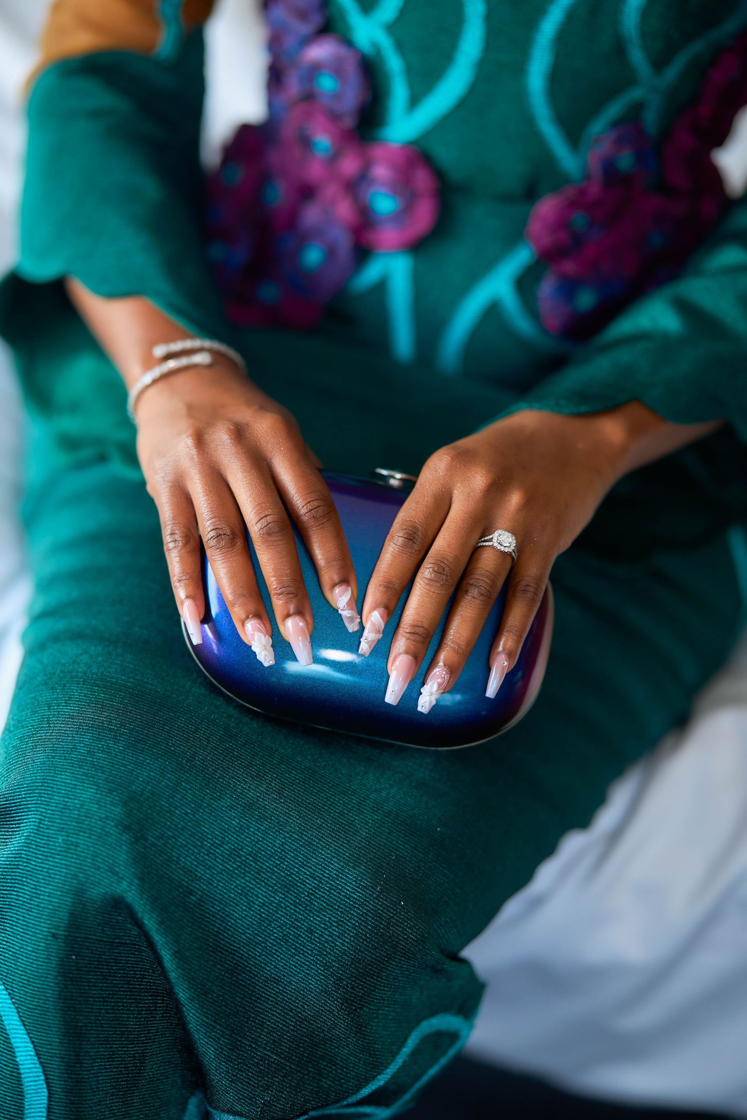 Woman in teal embroidered dress with long white nails and diamond ring holding a blue clutch purse.