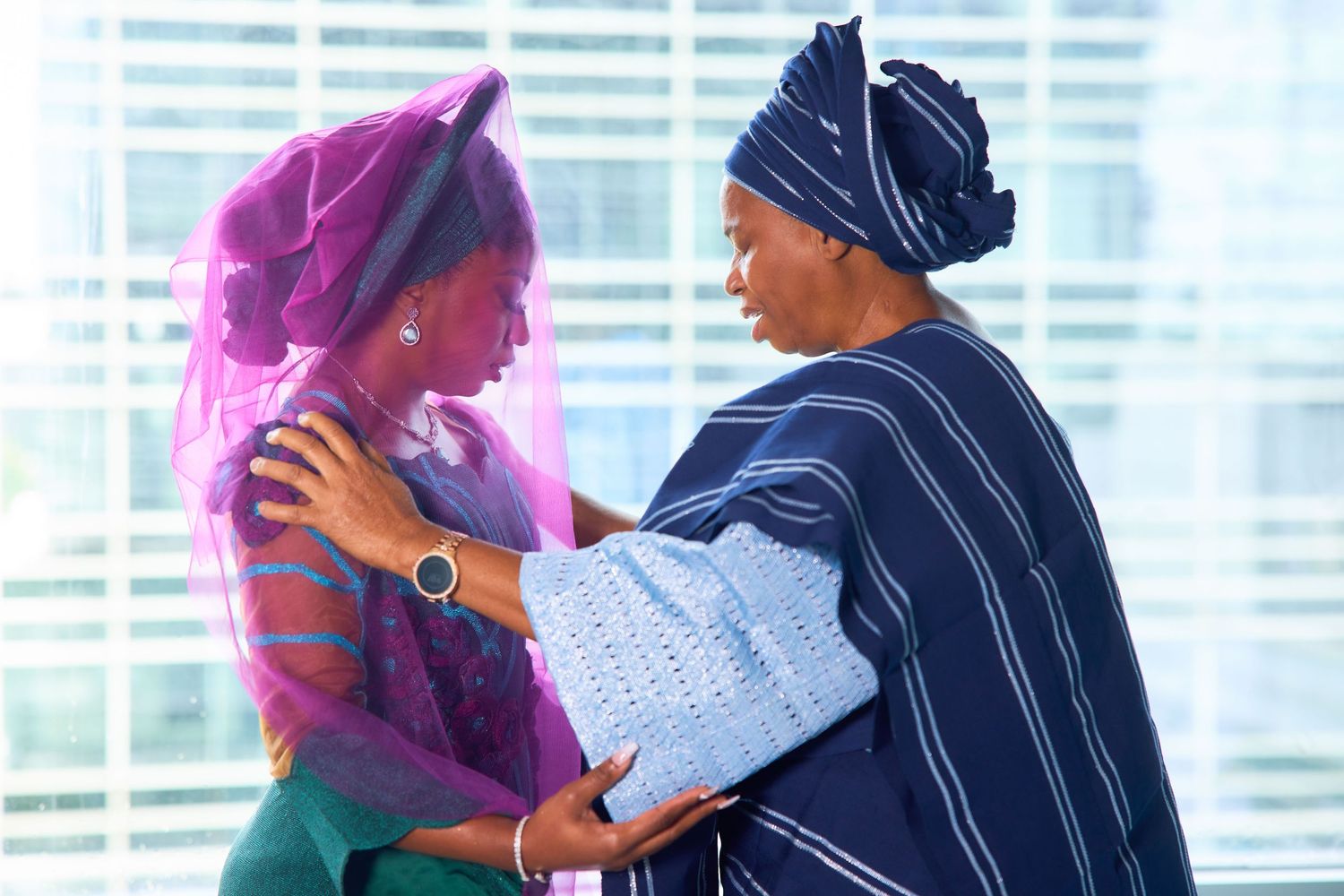 Two women in traditional African attire share a warm embrace, wearing vibrant blue and purple garments.